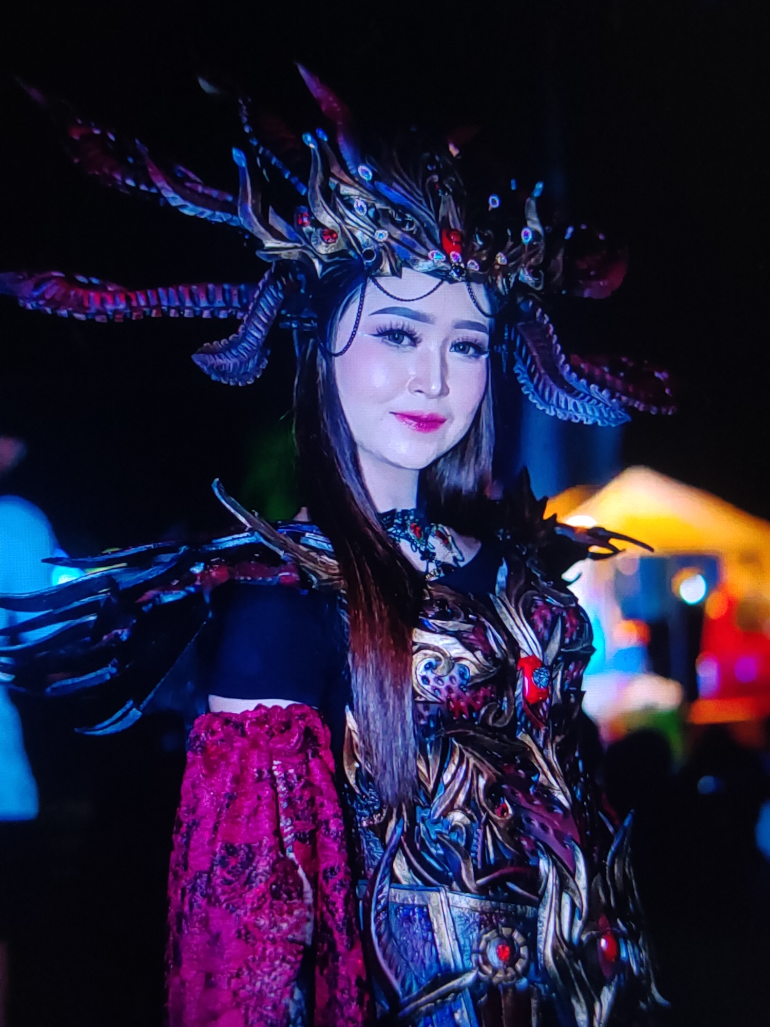 A woman in a detailed, ornate costume stands against a dark background with blurred lights in the distance. The costume features a large, elaborate headdress adorned with what appear to be feathers, horns, and jewels in shades of blue, purple, and red. She wears a matching chest piece with intricate gold and blue metallic details, accented with red jewels. Her shoulders are decorated with black, feather-like protrusions. A flowing, textured pink garment is visible on her left arm. The woman has long, dark hair and is wearing makeup, including pink lipstick and dramatic eyeliner. The overall impression is that of a performer in a parade, festival, or theatrical event. The location is identified as Unknown, Unknown.