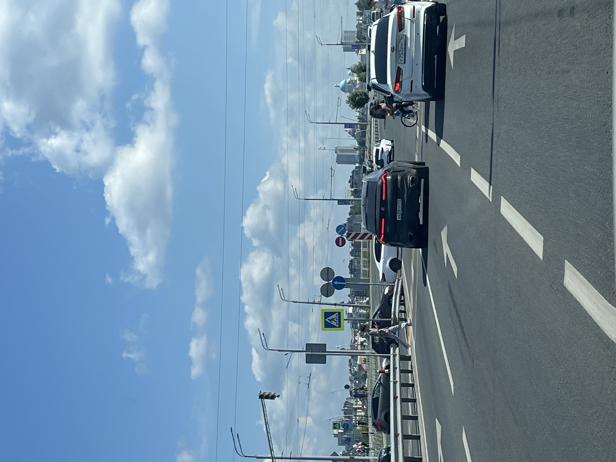 This is a vertical, eye-level, medium shot photograph taken from a moving vehicle, capturing a view of a multi-lane road in Kazan, Russia. The sky is predominantly blue with scattered white clouds. A pedestrian crossing is visible on the road.
In the foreground, the asphalt road surface is marked with white directional arrows. Several vehicles are present: a dark-colored BMW X6, a white Kia Optima, and a white sedan. A person is cycling on the road next to the white Kia.
In the middle ground, a pedestrian is walking across the pedestrian crossing, to the left of the dark-colored BMW. Other vehicles are visible on the road, including a dark-colored car and a white car. Traffic lights and street lamps are positioned along the side of the road. Pedestrian crossing signs, one of which is a yellow diamond with a blue pedestrian symbol, are also visible.
In the background, a cityscape with various buildings, including high-rise structures and a building with a blue dome, is discernible. Overhead power lines and traffic signals are also present. The overall environment appears to be a typical urban setting with infrastructure for transportation and city life. The lighting suggests it is daytime, likely mid-morning or afternoon, given the bright sky and clear visibility.
