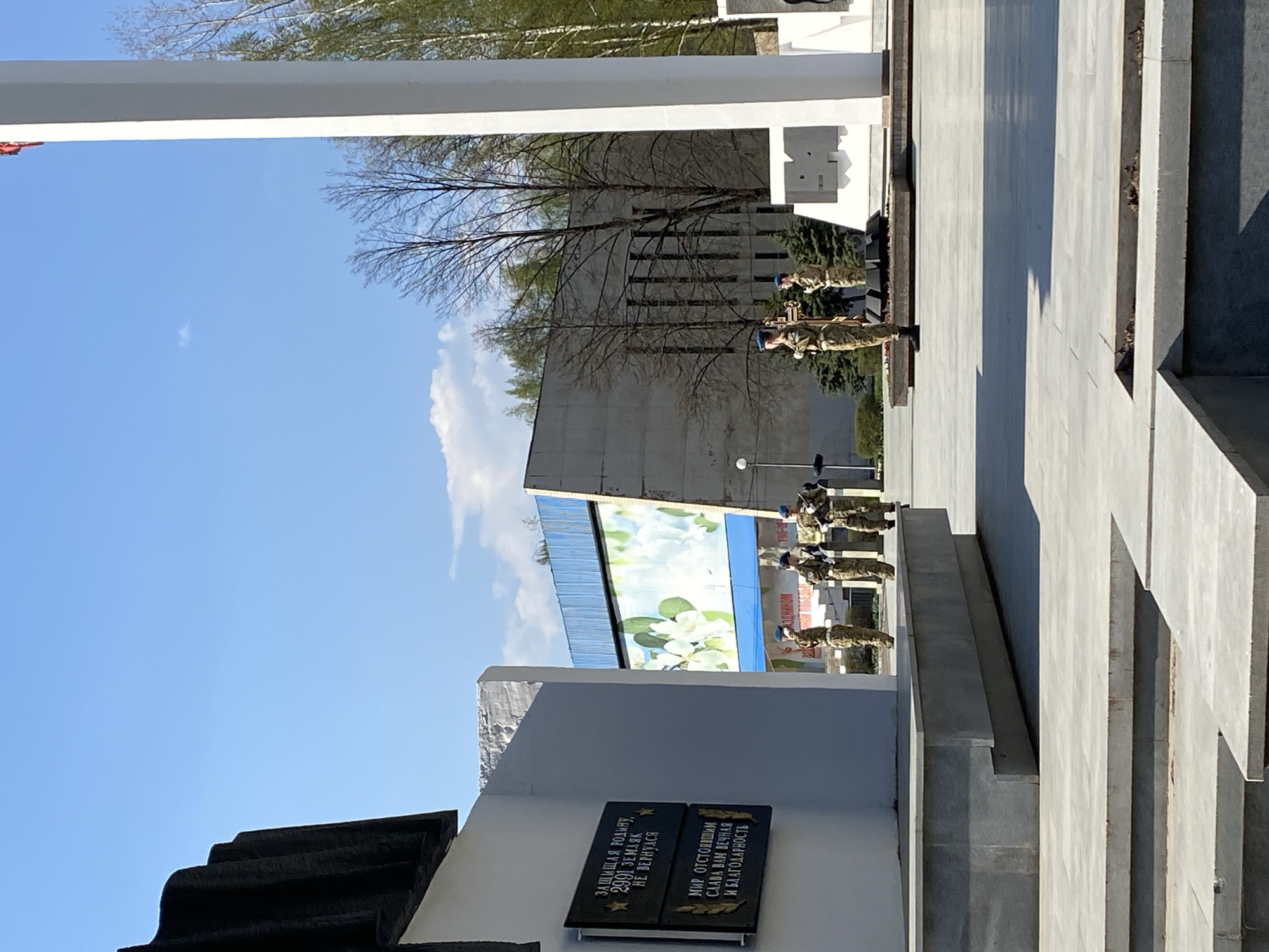 The image depicts a memorial complex in the city of Chaikovsky, Russia. Several soldiers in military uniforms and blue berets are standing at attention near a concrete structure. A plaque on the structure displays the inscription "ЗАЩИЩАЯ РОДИНУ, 2991 ЗЕМЛЯК НЕ ВЕРНУЛСЯ. МИР ОТСТОЯВШИМ СЛАВА ВАМ ВЕЧНАЯ И БЛАГОДАРНОСТЬ". The background includes buildings and trees under a blue sky.