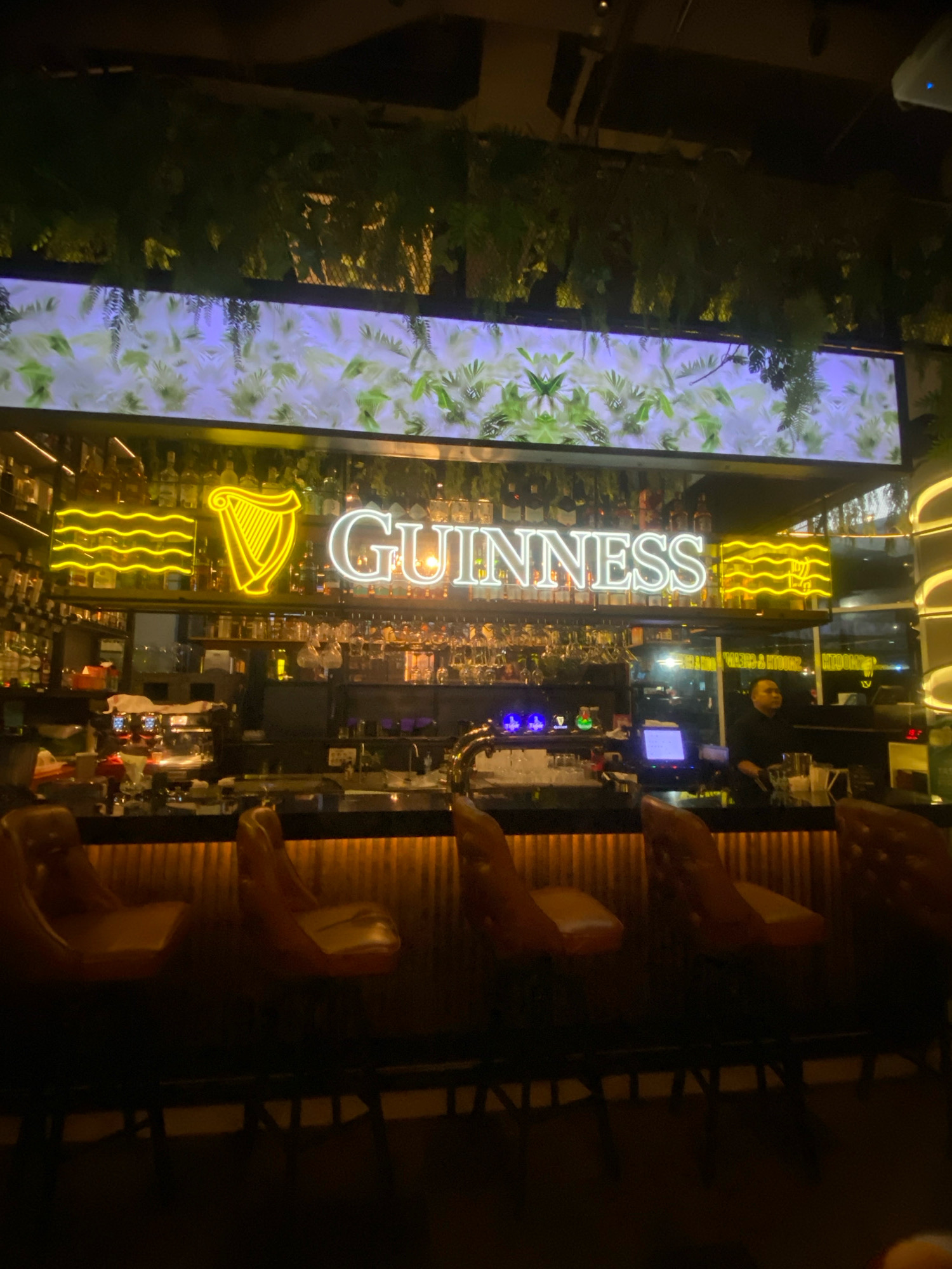 The image shows the interior of a dimly lit bar, identified as Guinness Store, located in Kuala Lumpur, Malaysia, likely in a hotel or upscale establishment. The central focus is a bar counter lined with tan leather bar stools. Behind the counter, a well-stocked bar features shelves filled with bottles of alcohol. Prominently displayed above the bar is a large, illuminated Guinness logo with a harp symbol. Neon signs in yellow and white spell out "GUINNESS" and also feature a yellow neon wave design and a harp. Various glassware hangs from racks above the bar. A bartender, wearing a black shirt, is visible behind the counter, attending to a customer or preparing drinks. The environment is sophisticated, with decorative plants hanging from above and subtle lighting creating a warm ambiance.