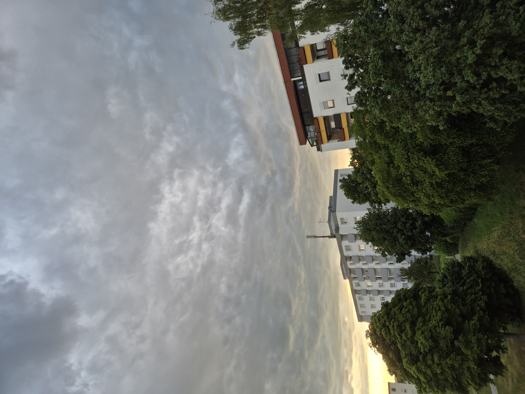The image displays an urban or suburban landscape under a heavily clouded sky during what appears to be late afternoon or early evening. The sky is completely covered by a dense layer of grey stratocumulus clouds, with a soft, diffused golden-yellow light emanating from the horizon on the far left, indicating the position of the sun behind the cloud cover.
Two main multi-story residential buildings are prominent in the midground. The building on the left is a long, white-facade structure with multiple windows and grey-railed balconies. A cellular antenna (telecommunications mast) is visible on its roof. To the right, a second apartment building, also white, features a distinct brown-red roofline and yellow panels on its balconies. A portion of a third white building is visible on the far left edge.
The foreground is heavily vegetated with various shades of green trees and bushes. A large, dark green, broad-canopied tree dominates the lower-left section of the frame. Numerous other trees and shrubs are scattered throughout the midground, partially obscuring the base of the buildings. A tall, slender tree with light green foliage stands on the far right. A grassy bank or slope extends across the lower-middle foreground. No individuals or vehicles are present. The scene depicts a quiet residential environment in an unspecified city.