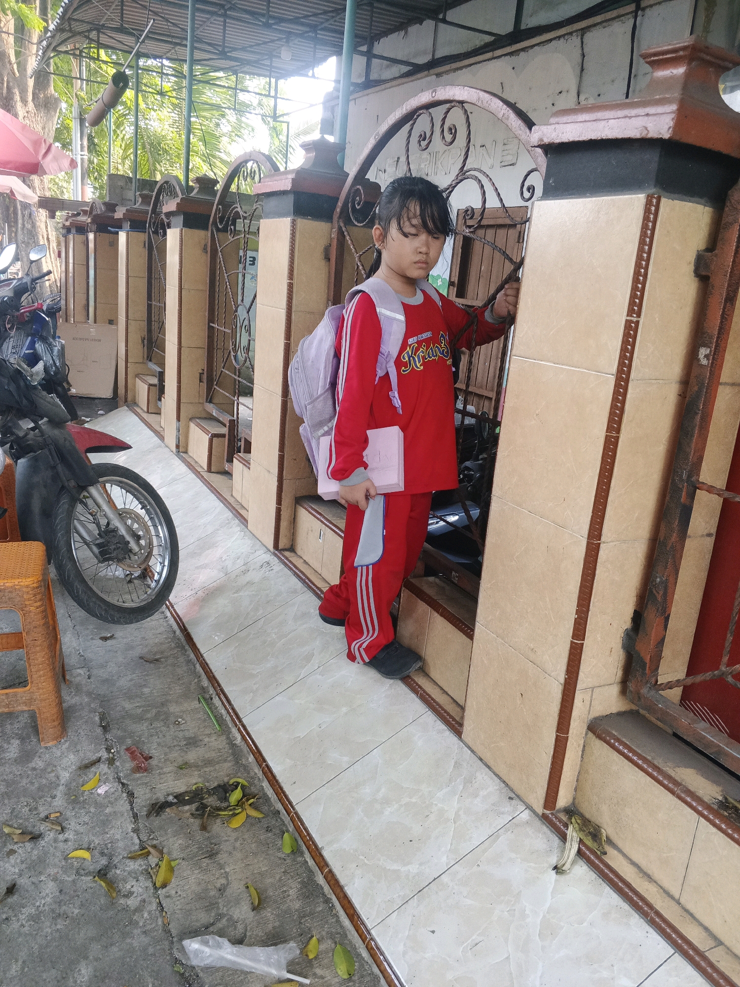 A young girl wearing a red tracksuit and a light purple backpack stands by a brown and beige tiled wall with an ornate metal gate. She is holding a pink object, possibly a book or tablet, in her right hand and gripping the metal gate with her left. The ground is tiled with a light-colored pattern. To the left of the girl, a motorcycle is parked next to an orange plastic stool on a concrete surface littered with fallen leaves and debris. The environment appears to be an outdoor area, possibly a school or public building in Krian, Indonesia, given the architectural style and the presence of a motorcycle. The lighting suggests it is daytime, with some sunlight filtering through the overhead structure.