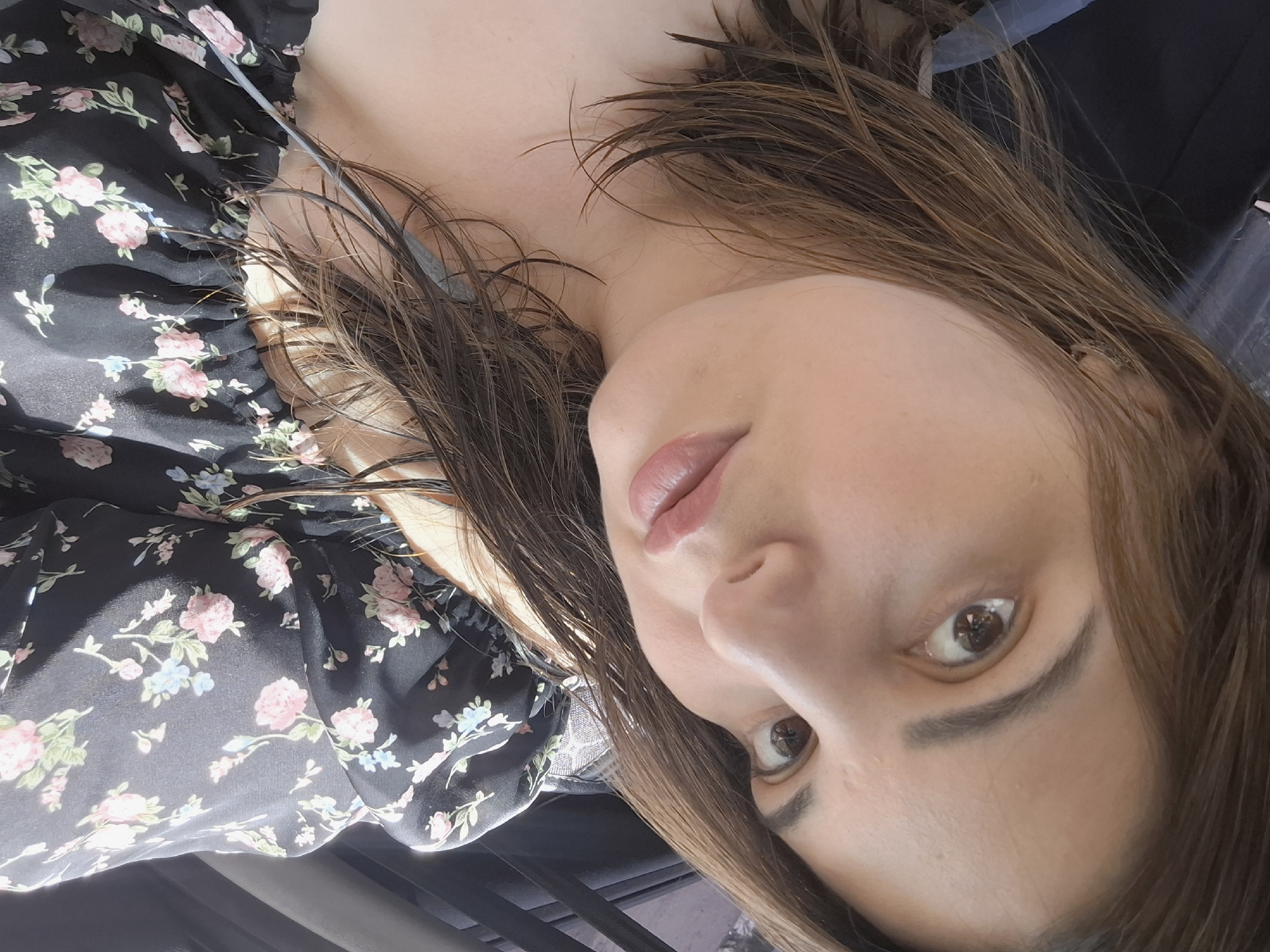 An overhead, close-up shot captures a young woman with long, brown hair pulled back as she looks up and to the right. She wears a black, off-the-shoulder top with a floral pattern of pink and blue flowers. Her makeup includes a subtle pink lip gloss. The background is dark and out of focus, suggesting an indoor setting with some natural light filtering in. No specific location in Chaikovsky, Russia, is discernible from the image.