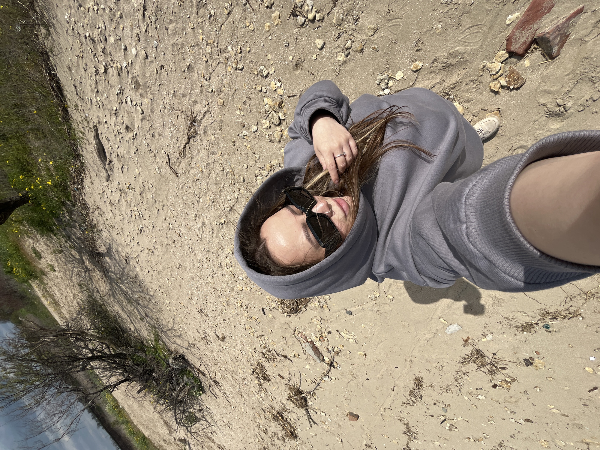 A young woman wearing a gray hooded sweatshirt and sunglasses is lying on a sandy surface. She has brown hair and is wearing a ring on her left hand. The ground around her is sandy, with small stones, shells, and debris scattered about. There are also fragments of brick or tile and dried plant matter. To the left, there is a patch of green vegetation and a bare tree. The scene suggests a location near a beach or shoreline. The location is Port-Katon, Russia.