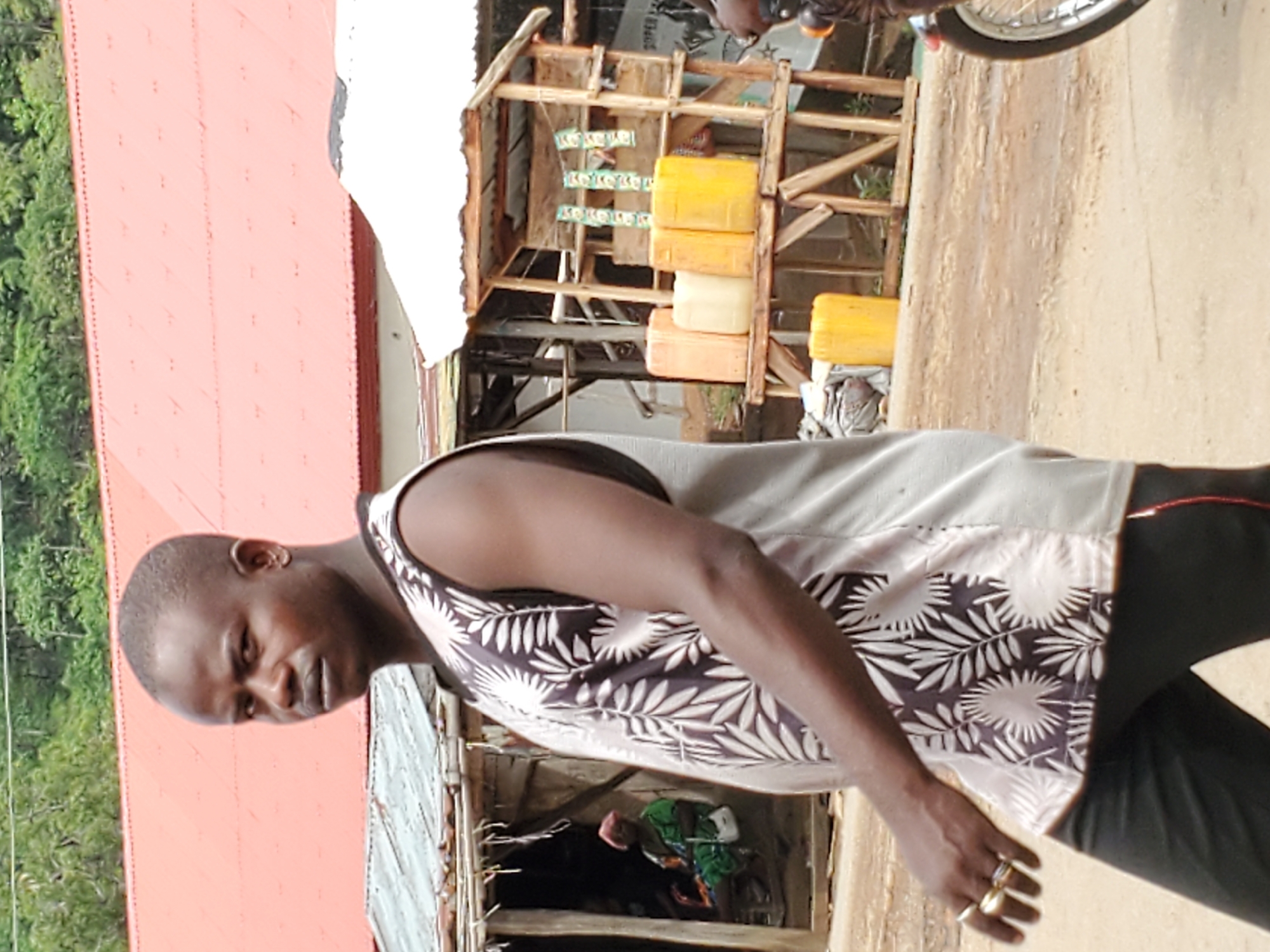A man stands outdoors in Mararaba, Nigeria, next to a building with an orange roof and a small wooden structure displaying yellow jerrycans and price signs. A motorcycle is also visible. The man wears a sleeveless shirt with a leaf pattern and dark pants. He has gold rings on his fingers. The background includes trees and foliage.