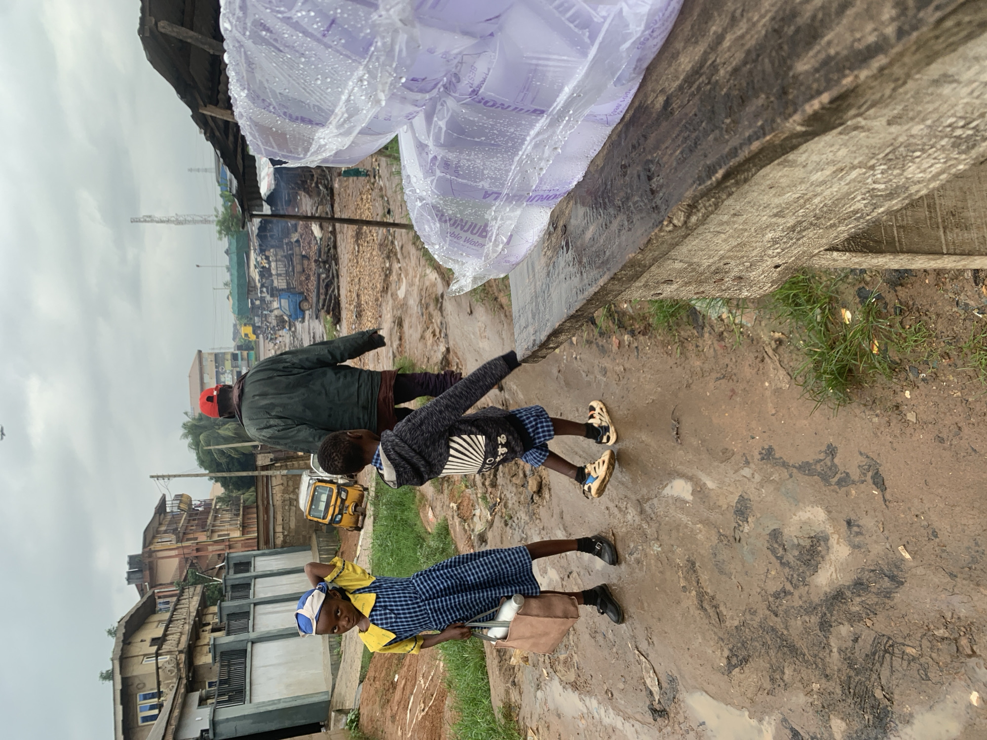 Two children, a girl and a boy, stand on a muddy, uneven path in what appears to be an outdoor market or settlement area. The girl, on the left, wears a blue and white checkered dress over a yellow collared shirt and a blue and white head wrap. She carries a light brown bag in her left hand and holds her right hand to her head. The boy, to her right, wears a dark hooded jacket with a graphic design on the back and blue and white checkered shorts. He walks with his right foot forward, arms slightly bent. Behind them, a man in a green jacket and a red cap walks away from the camera. In the background, there are multi-story buildings, a yellow tricycle, and various market stalls with corrugated metal roofs. The ground is wet and muddy, indicating recent rain. To the right of the image, a stack of what appears to be packaged water pouches is visible, partially covered by plastic sheeting and resting on a weathered wooden bench. The sky is overcast.