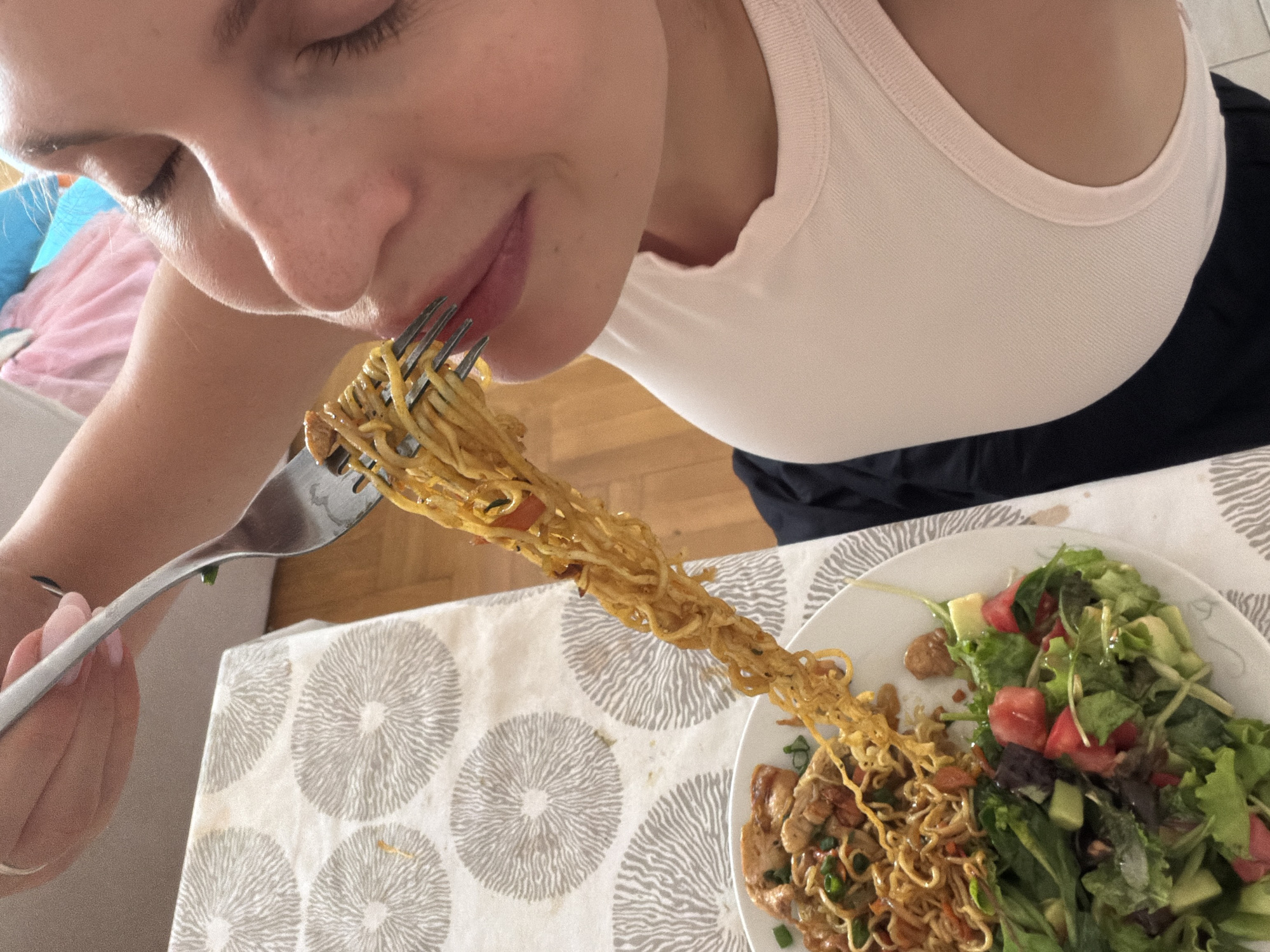 In Budva, Montenegro, a close-up shot captures a person eating noodles from a fork. The person's face is partially visible, with their eyes closed and lips slightly parted, as they bring a forkful of noodles to their mouth. The noodles appear to be stir-fried, with bits of vegetables and meat mixed in. A white plate with a salad and more noodles is visible in the foreground, and a patterned tablecloth covers the table. The scene suggests a casual meal in a home setting.