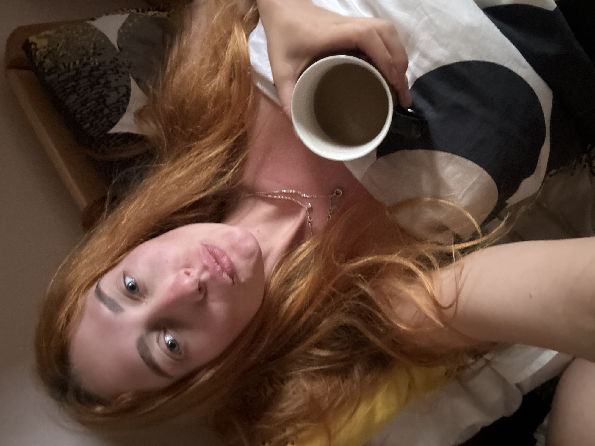 A single young adult female subject, estimated to be in her twenties, is captured in a selfie-style photograph. She lies partially reclined in a bed, facing the camera with her head angled slightly to her left. Her long, wavy, reddish-orange hair is spread across a white pillow on the left and over her shoulders and chest. Her fair skin shows a subtle flush across her cheeks and nose. Her eyes are open and light-colored. Her lips are pursed into a slight pout. She wears a delicate silver-colored chain necklace with small, reflective charms around her neck.
Her right hand (viewer's left) grasps a black cylindrical mug with a white interior rim, which contains a light brown liquid consistent with coffee or tea. The subject's upper body is partially obscured by white bedding, which features a prominent abstract pattern of large black crescent moon or partial circular shapes against the white fabric. A yellow pillow is visible behind her head on the left side of the frame. In the upper right background, behind her hair, a wooden headboard or piece of furniture is discernible, alongside a pillow or fabric item adorned with a dark green/grey pattern accented by lighter, abstract shapes. The scene is illuminated by soft, ambient light, typical of an indoor residential setting in the city of Budva, Montenegro.