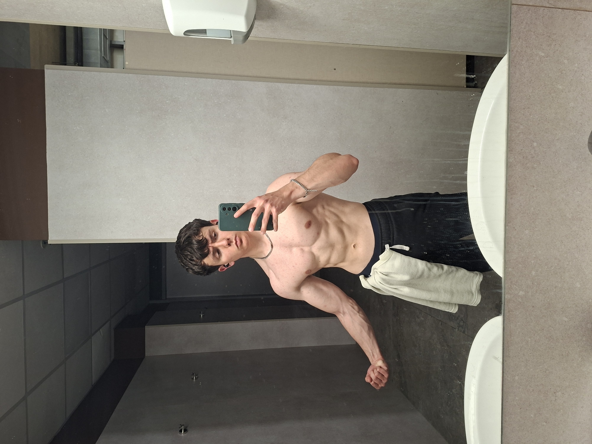 A shirtless young man stands in what appears to be a public bathroom. He holds a smartphone in his right hand, using it to take a selfie in a mirror. He is wearing dark-colored sweatpants and a silver chain around his neck. A white towel is tucked into the waistband of his pants. The bathroom features light-colored wall tiles, a white sink, and a stainless steel hand dryer. The ceiling is dark and tiled. There is no indication in the image to suggest this is located in the city of Plovdiv, Bulgaria.