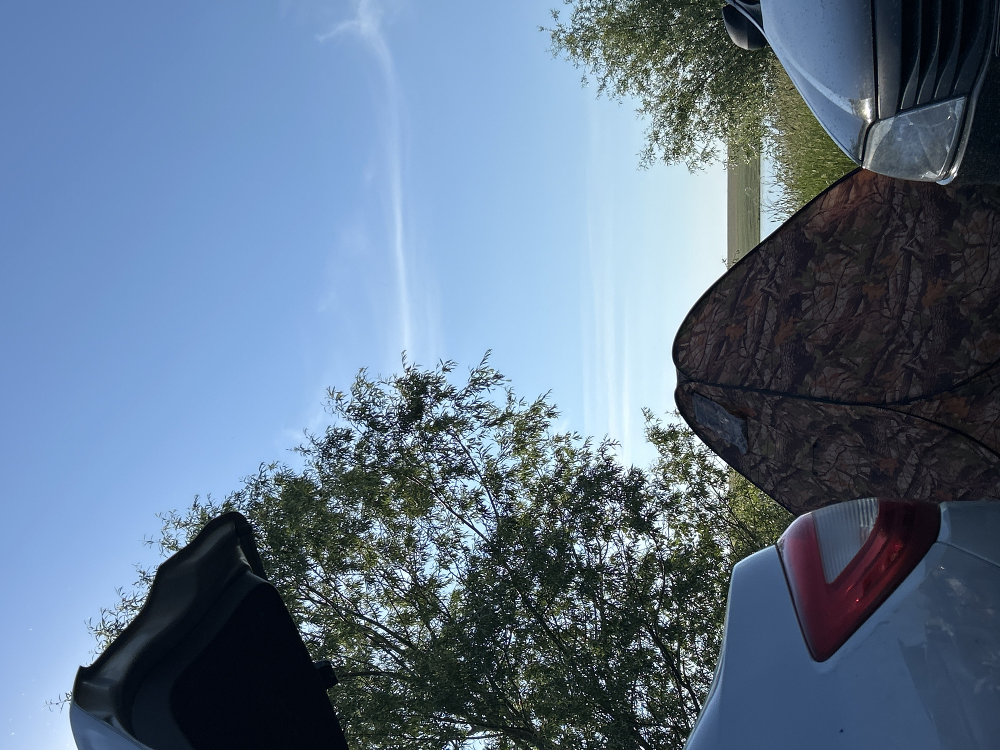The image depicts an outdoor scene with two cars, a camouflage tent, and trees against a bright blue sky with wispy clouds. On the left, the rear of a light-colored car is visible, showing its taillight. In the center, a camouflage tent is set up, partially obscured by the cars and foliage. On the right, the front of a dark-colored car is visible. Trees with green foliage are present on the left and right sides of the frame. In the background, a body of water is visible, with a grassy field beyond it, leading to a distant horizon under the expansive sky. The setting appears to be a natural environment, possibly a campsite or a roadside stop in a rural or natural area. The specific location in Matrayevo, Russia, is not visually identifiable from the image alone.