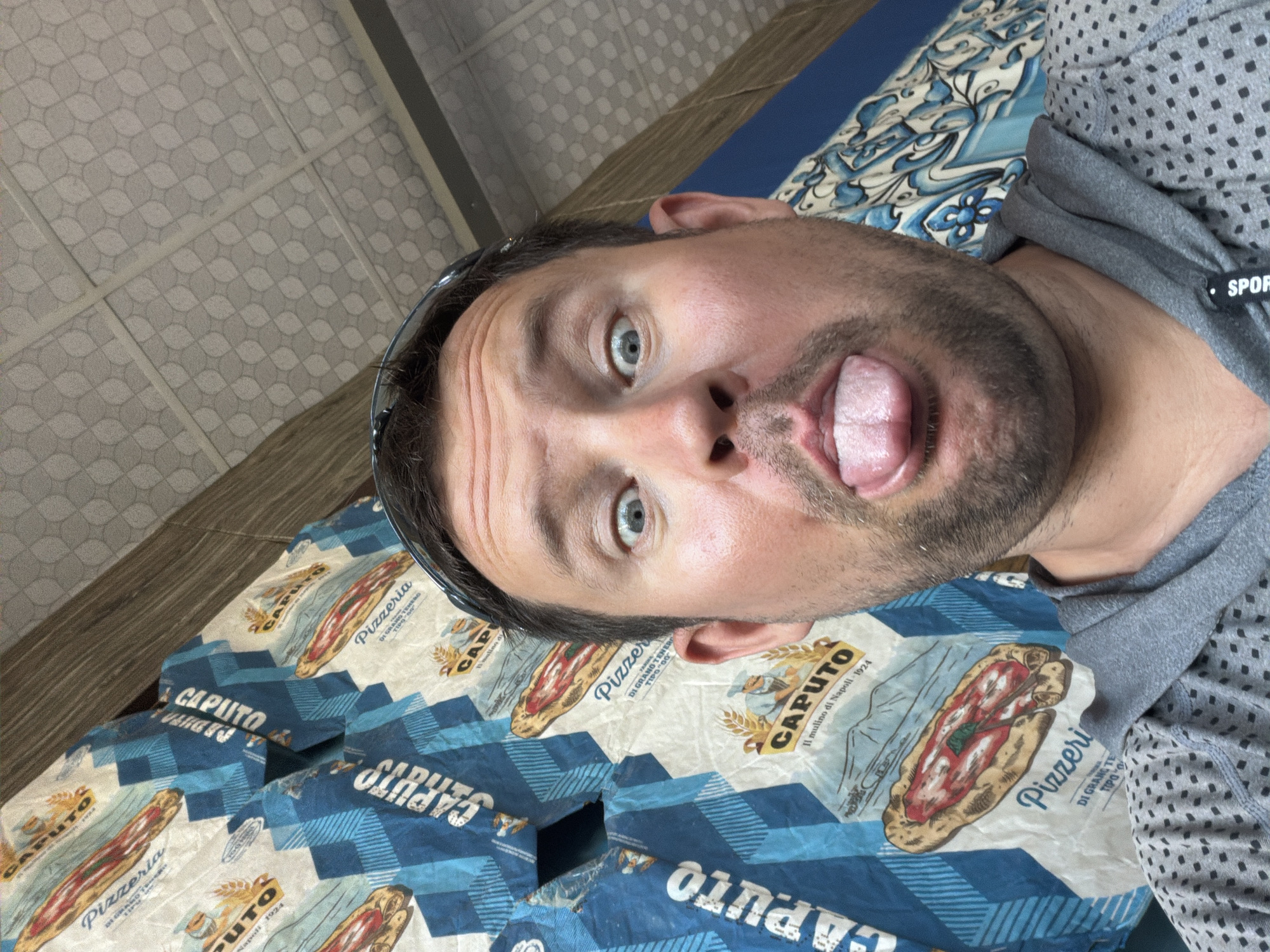 A man is taking a selfie with his tongue sticking out. He is wearing a grey hooded sweatshirt with a black geometric pattern. He has short brown hair and a beard. The background consists of stacks of flour bags with a blue and white checkered pattern and images of pizza. The ceiling above him has a patterned design. The overall impression is informal and playful. There is no indication of Baan Bo Phut, Thailand in the image.