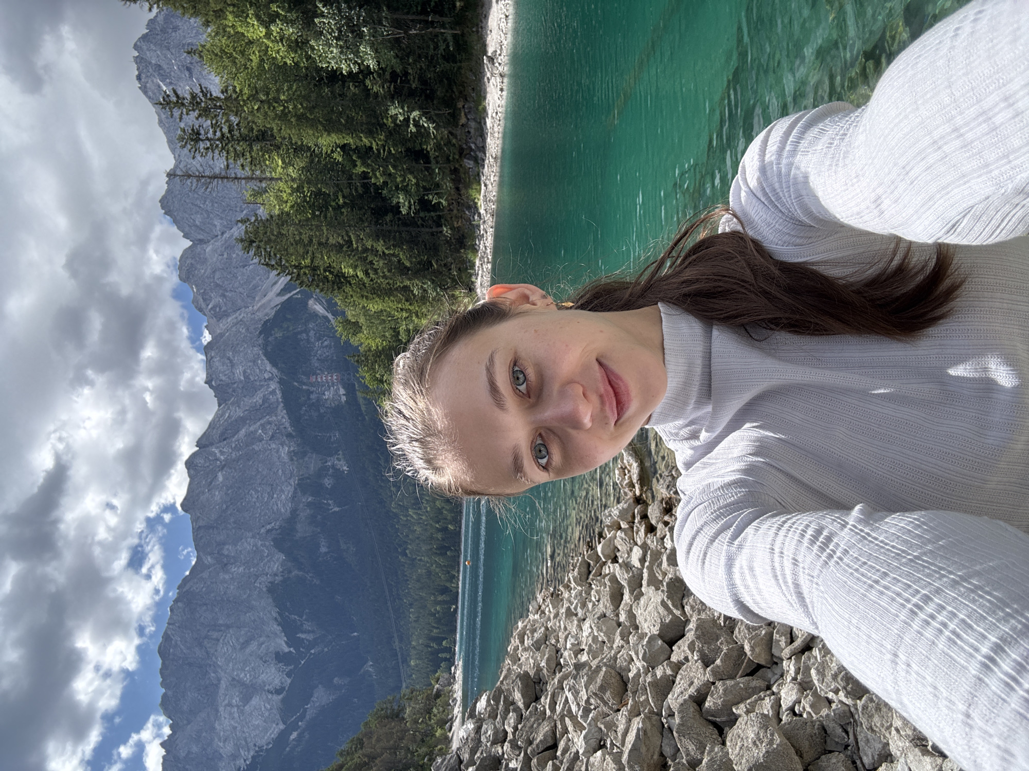 A young woman is taking a selfie in front of a vibrant turquoise lake with majestic mountains in the background. The lake is situated in the Bavarian Alps, near Grainau, Germany, specifically in the area of the Eibsee, identifiable by its unique color and surrounding rocky shoreline. The woman is wearing a light gray, high-necked sweater. Her hair is pulled back into a ponytail, and she has a friendly smile. The background features a dense forest of evergreen trees climbing the lower slopes of the mountains, which are jagged and appear to be composed of gray rock. The sky is partly cloudy with dramatic cloud formations. The foreground consists of a shoreline covered in light-colored rocks.