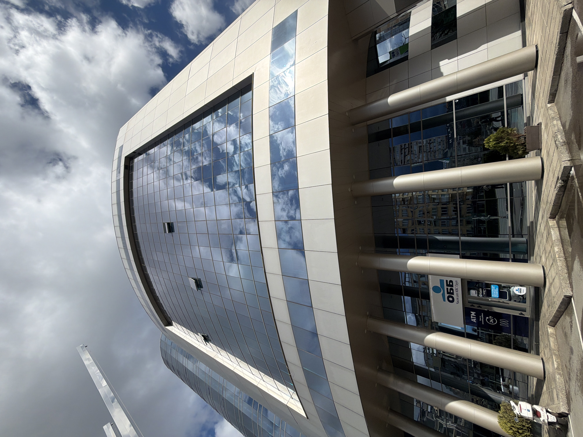 The image depicts a modern office building in Sofia, Bulgaria. The structure features a curved, glass-clad facade that reflects the sky and clouds. Cylindrical columns support the building's entrance, where an OBB bank branch is located. The surrounding environment includes a sidewalk and street. The building's exterior is predominantly white and gray, with glass panels forming a grid-like pattern.