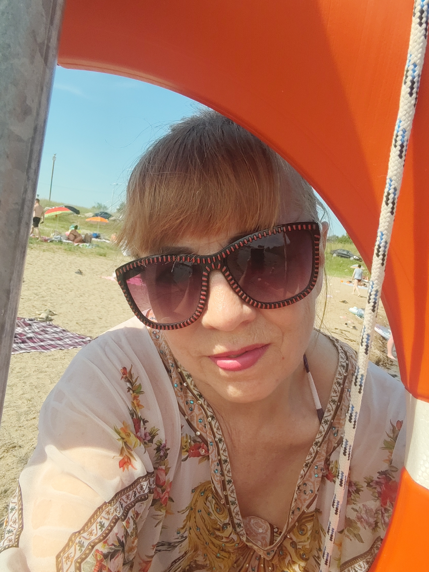 A woman is taking a selfie at a beach. She is wearing large, decorative sunglasses and a sheer, floral print cover-up over a bikini top. The woman has blonde hair and is looking directly at the camera. The background shows a sandy beach with a few people in the distance, beach umbrellas, and the blue sky. A part of an orange life preserver with a rope is visible on the right side of the frame, and a grey pole is on the left. The overall scene suggests a sunny day at a recreational area. There is no specific indication of the city of Rovnoe, Russia in the image.