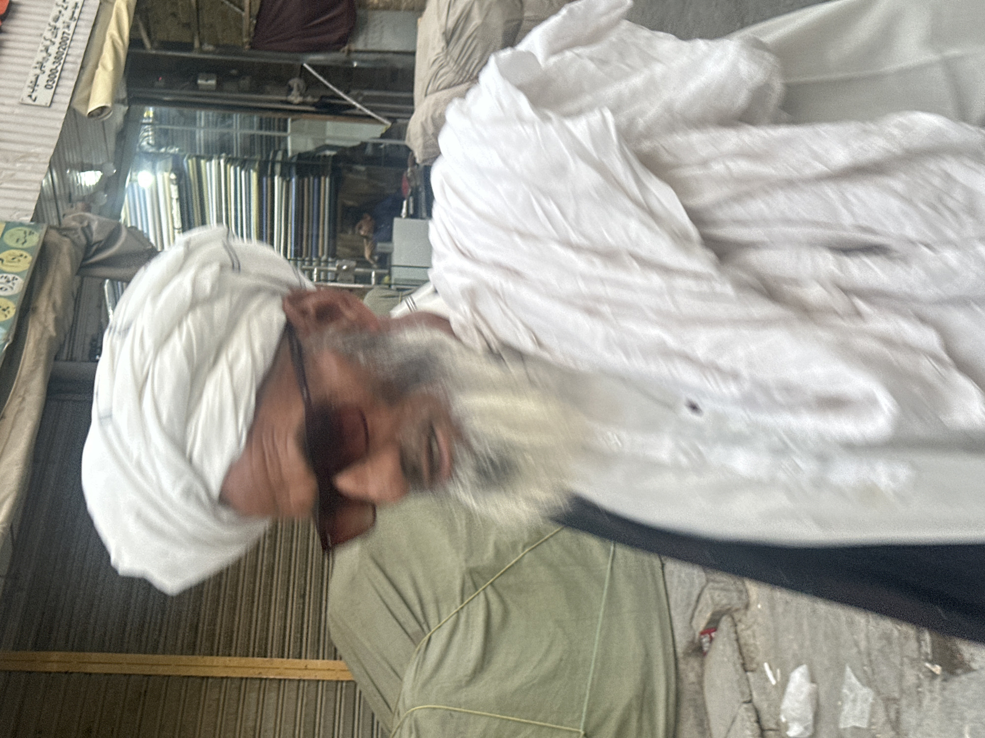 A medium shot shows a man in a white turban and white clothing walking outdoors, possibly in a marketplace. He has a beard and is wearing sunglasses. The background is blurred but shows what appears to be shops with shelves of fabric and metal shutters. A green tarp-covered object is visible to the left. The scene is outdoors and appears to be a street or alleyway. The lighting is natural daylight.