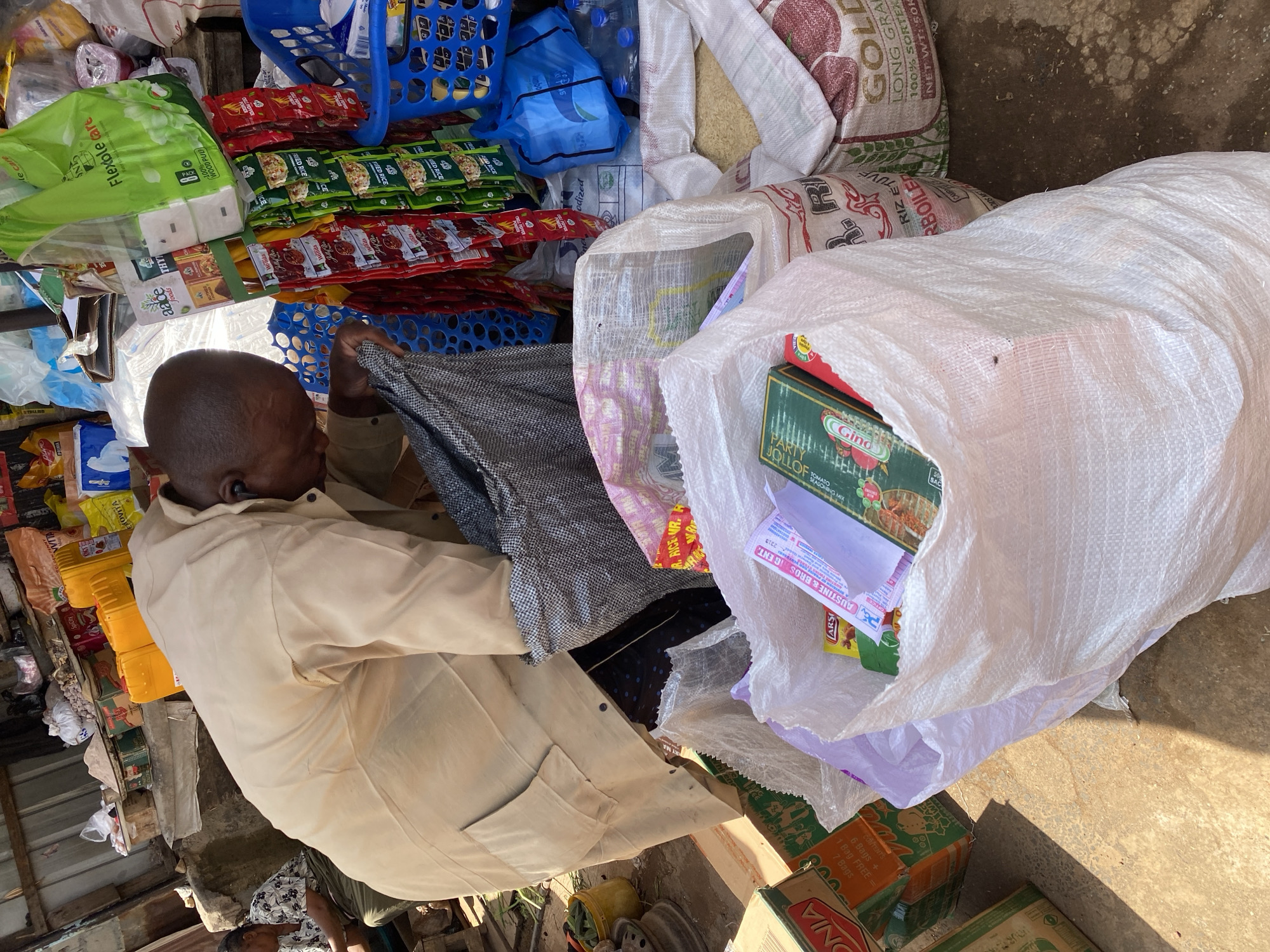 This media file shows a market scene in Jos, Nigeria, with a focus on a stall selling various food items and household goods. The primary subject is a man, likely a vendor, in a light-colored smock, who is partially visible from the side, looking downwards. He appears to be arranging or handling some goods.
The stall is densely packed with a variety of products. Large sacks of rice, some white and some with red and gold packaging, are prominent. Several smaller packets of what appears to be seasonings or spices, with red and green packaging, are neatly arranged. There are also bags of flour or similar dry goods. Among the items are boxes of "Gino Jollof Tomato Seasoning Mix." Blue plastic baskets, some filled with water bottles and other items, are also visible. In the foreground, a large white woven sack is open, revealing more packaged goods. The ground appears to be dirt or concrete, characteristic of an outdoor market.
The lighting suggests it is daytime, with sunlight casting shadows, indicating a warm climate. The overall impression is of a busy, perhaps bustling, market environment. The man's posture and the arrangement of goods imply an ongoing commercial transaction or preparation for one. There is no clear indication of the specific time of day or weather conditions beyond the presence of sunlight. The visible text on the products includes brand names like "Gino," "FlexiCare," and "Gold," along with indications of product type such as "FRIED RICE," "TOMATO SEASONING MIX," and "LONG GRAIN."