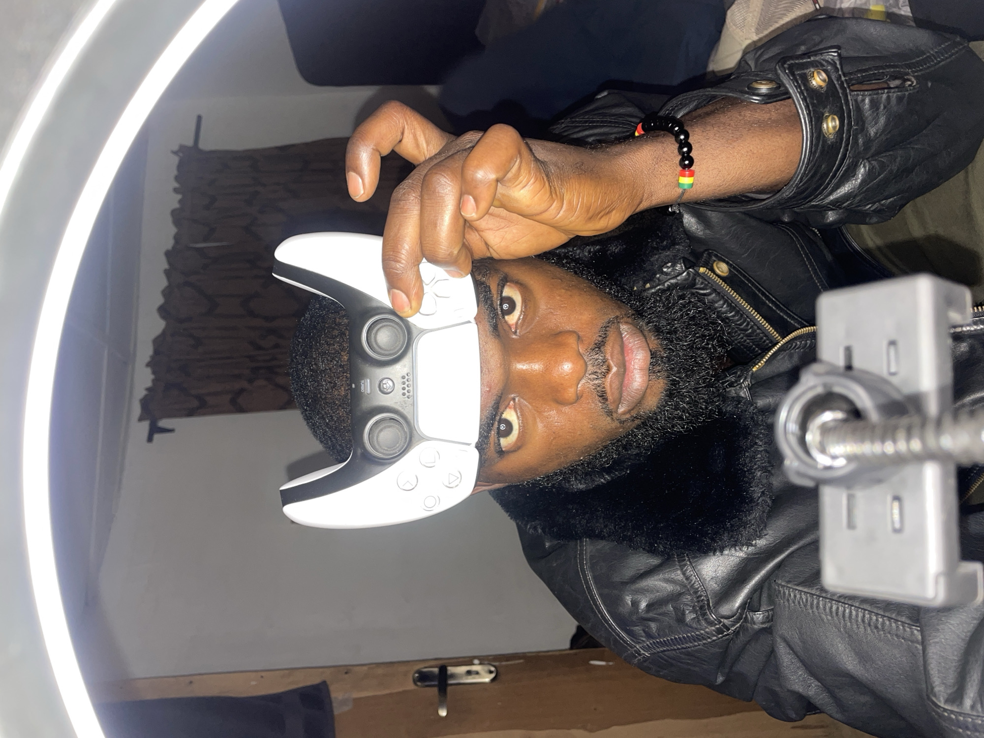 A dark-skinned man with a beard is holding a white PlayStation 5 controller over his right eye. He is wearing a black leather jacket with a fur collar and a black and red-green-yellow beaded bracelet on his right wrist. He appears to be in front of a ring light. A silver phone mount is positioned to his right. The location is not discernible and therefore cannot be determined to be Jos, Nigeria.