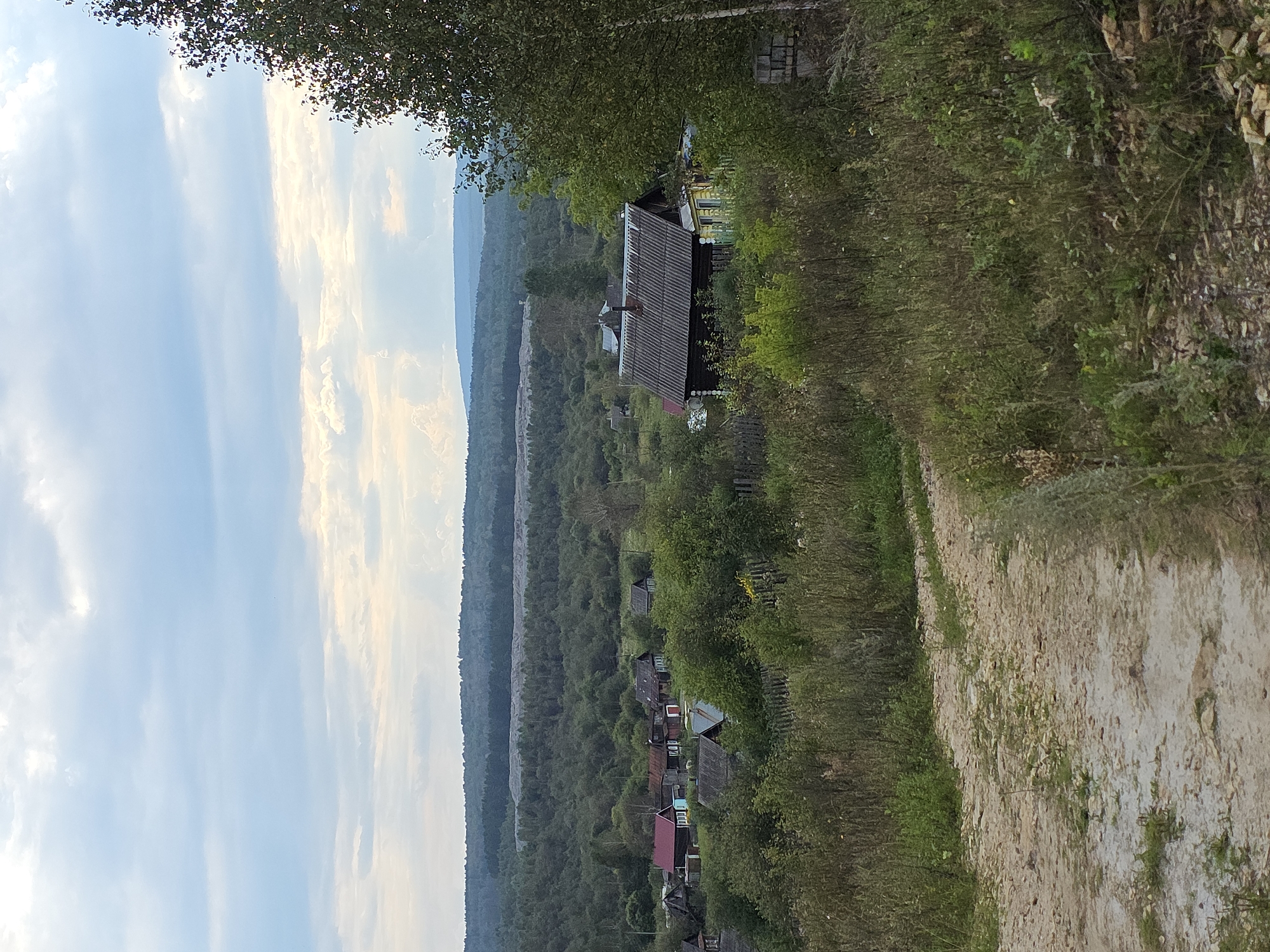 This is a daytime, medium-long shot photograph taken from an elevated position overlooking a rural village. In the foreground, a dirt road curves from the bottom left towards the center of the frame, bordered by overgrown grass and vegetation. A few scattered rocks are visible on the road and surrounding terrain.
The middle ground features a cluster of rustic houses, mostly single-story structures with pitched roofs made of slate or corrugated metal. Some houses have visible wooden walls, while others are painted in muted colors like red, blue, and yellow. Fences made of wood are present around some of the properties. The houses are nestled among dense green trees and bushes, creating a somewhat overgrown appearance.
In the background, a vast expanse of dark green forest stretches across the horizon. Beyond the forest, a lighter, flat-topped hill or mountain range is visible in the far distance, under a cloudy sky. The sky is predominantly light blue with scattered white and gray clouds, some illuminated by sunlight. The overall scene depicts a tranquil, rural setting in the city of Magnitka, Russia, characterized by natural landscapes and traditional village architecture.