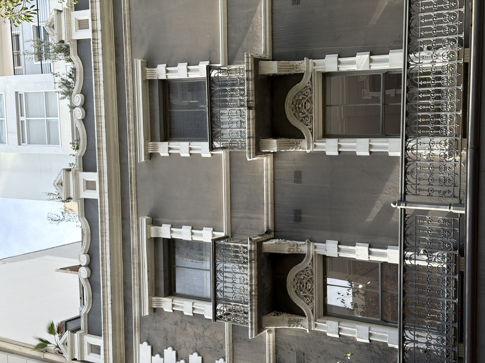 The image shows the facade of a building in Cape Town, South Africa. The building has a grey exterior with white window frames and ornamental details. Two rows of windows are visible, with decorative balconies attached to some of them. A section of a black wrought-iron railing runs along the side of one of the windows. Another white building is to the left.