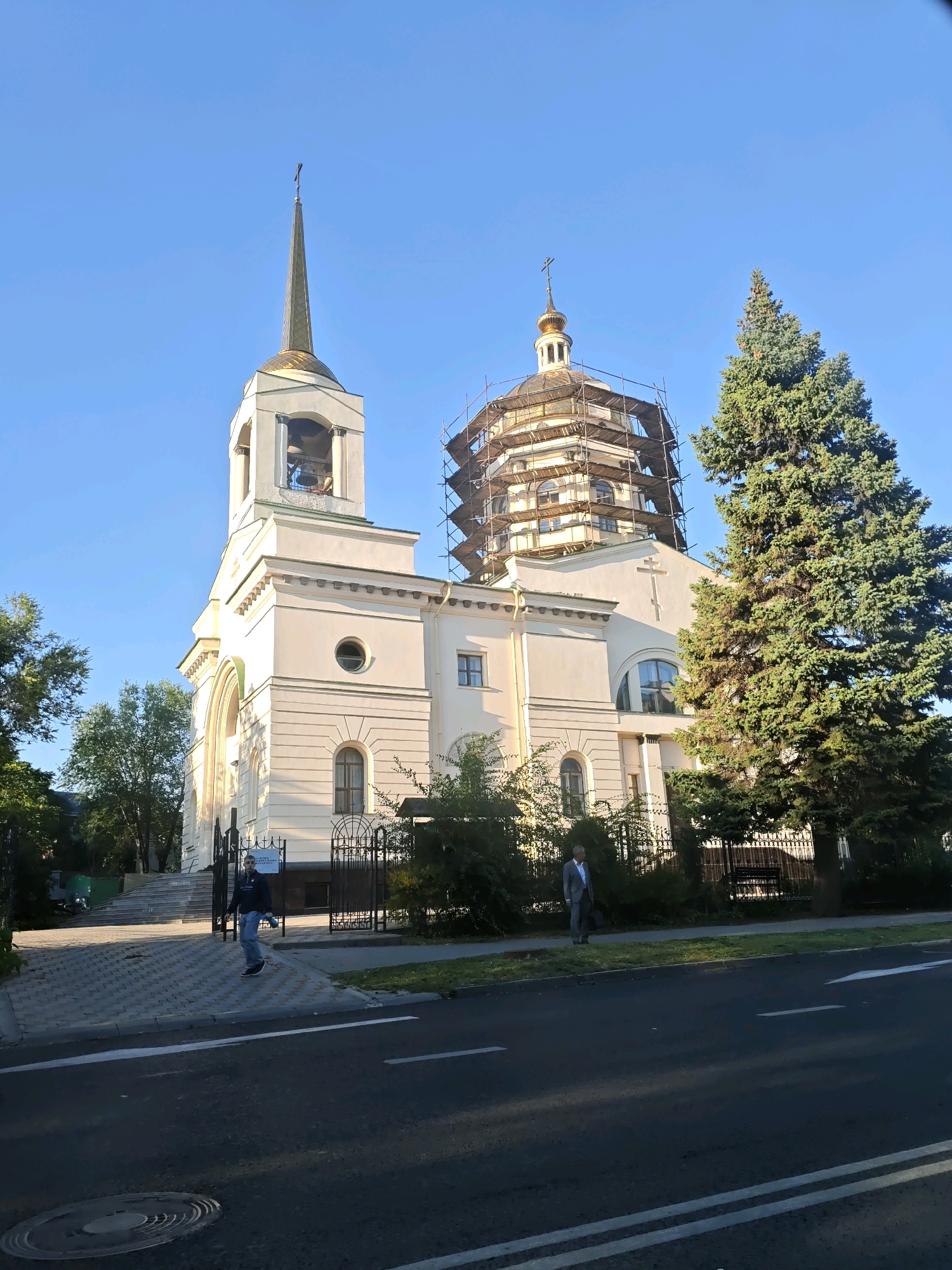 The image shows a cream-colored church with a tall, slender spire topped by a cross, and a larger dome covered in scaffolding. A large green conifer tree stands to the right of the church. In the foreground, there is a paved walkway and a street with white lane markings. Two individuals are visible: one man in a dark jacket and jeans walking on the walkway, and another man in a suit walking on the grass verge by the street. The sky is clear and blue, suggesting it is daytime. The architecture suggests it could be an Eastern Orthodox church.
This is a view of the Ascension Cathedral in Rostov-on-Don, Russia.