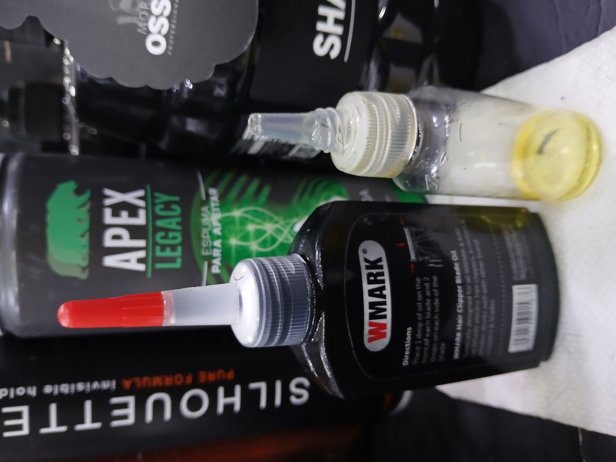 The image displays an assortment of grooming and hair care products. These items are clustered together on a white surface.
A bottle of "SILHOUETTE" hair spray with the tagline "PURE FORMULA Invisible Hold" forms a base layer. Adjacent to it, an "APEX LEGACY" bottle is positioned, labeled as shaving foam ("ESPUMA PARA APEITAR"). Overlapping these items is a black bottle labeled "WMARK Hair Clipper Blade Oil" with usage instructions. Next to it is a clear bottle containing a white liquid and a yellow top.
The text also indicates that the WMARK product is "Specially designed for all WMARK & ANDIS", indicating the types of hair clippers and blades the oil is compatible with.