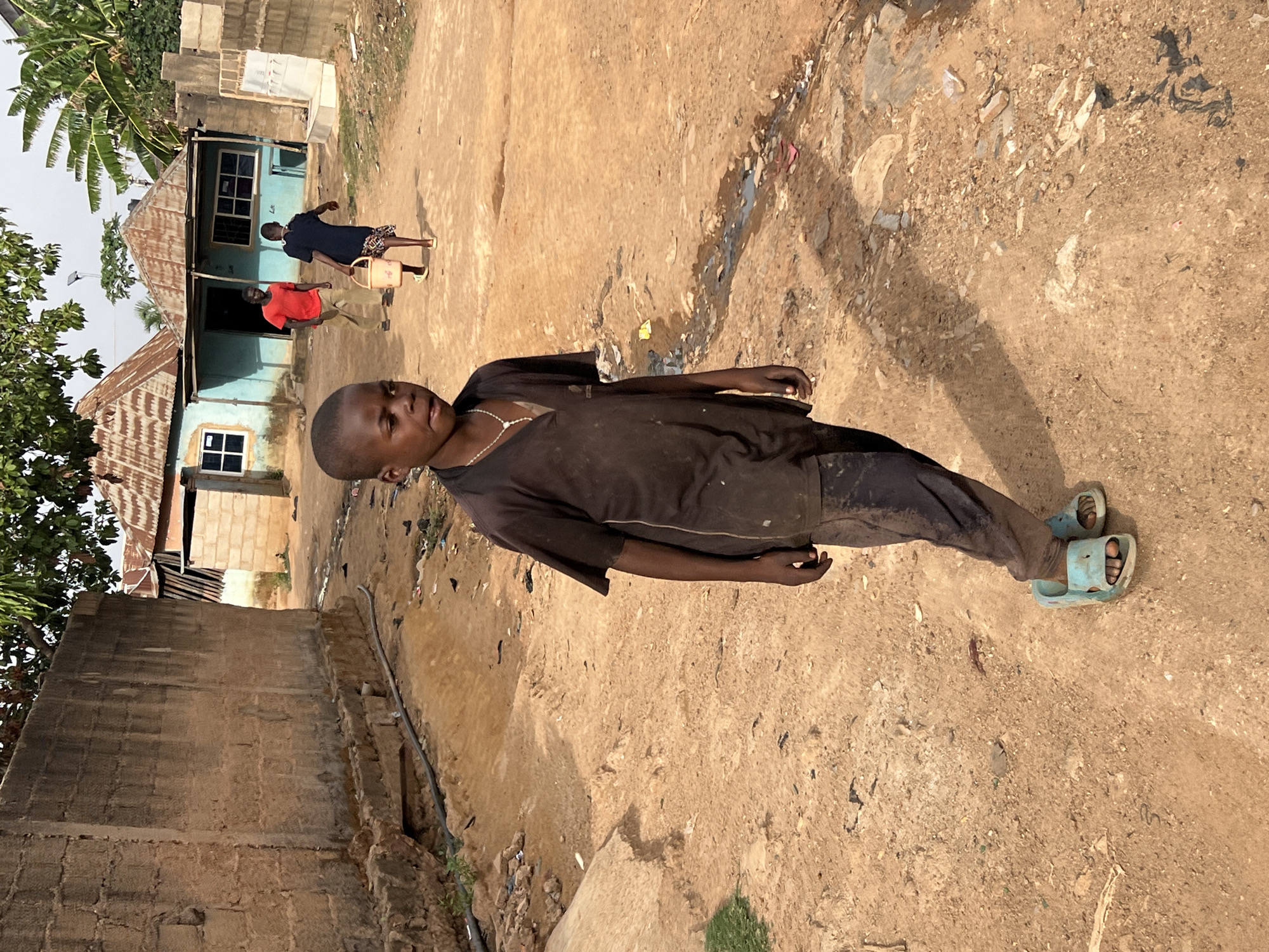A young boy stands in the foreground on a dirt road in Gura Top, Nigeria. He is wearing a dark brown shirt, dark pants, light blue sandals, and a necklace. In the background, two people are walking in front of a light blue house with a rusted metal roof. One person is wearing a red shirt and the other is wearing a dark blue shirt and carrying a bucket. The road is littered with trash. There is a brick wall on the left side of the image and trees and plants on the left and in the background.