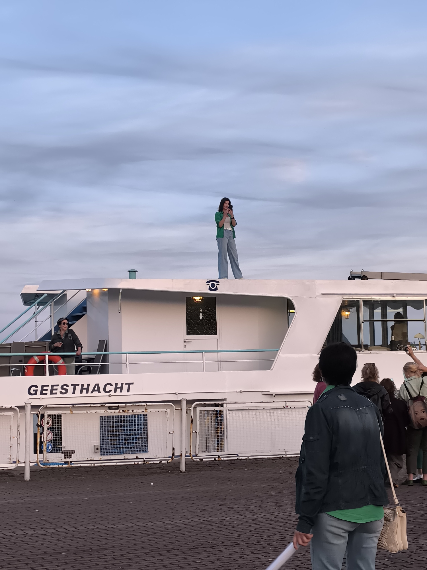 The image captures a group of people gathered near a white boat named "GEESTHACHT," docked at a waterfront location. The boat has a white upper deck where two individuals are standing. One person, dressed in a green jacket and light blue pants, is standing on the top of the boat, holding a phone and appearing to take a picture. Another person, wearing a dark jacket, sunglasses, and a cap, is leaning on the railing of the boat's deck.
In the foreground, several people are visible from behind, facing the boat. One individual in the immediate foreground wears a dark denim jacket over a green shirt and light blue pants, carrying a light-colored purse and holding a white object, possibly a rolled-up poster or banner. The ground is paved with dark cobblestones. The sky is overcast with soft, diffused clouds. The overall scene suggests an event or gathering taking place at the harbor. The presence of a life preserver on the boat and the signage on the lower deck indicate it is a functional vessel. The signage on the boat, "GEESTHACHT," does not directly provide a location, but the question specifies Neringa, Lithuania as the context.