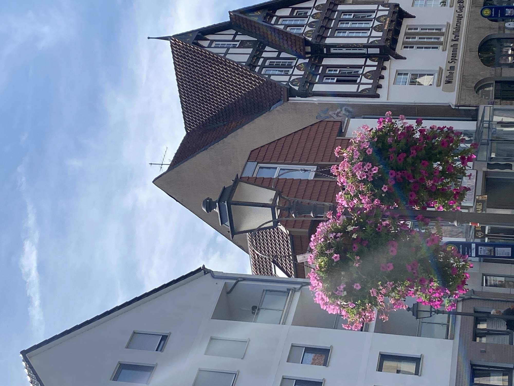 This image depicts a street scene in the city of Bückeburg, Germany, on a bright, sunny day with a partly cloudy sky. The foreground is dominated by two large hanging baskets overflowing with vibrant pink petunias, suspended from a black lamppost. The lamppost itself is a traditional design with a glass-paned lantern.
Behind the flowers and lamppost, a modern white building with multiple windows and balconies is visible on the left. To the right of this building, an older, traditional German half-timbered building with a steeply pitched, dark tiled roof stands out. This building features decorative dark wood beams against a white facade and several multi-paned windows. A sign above the arched entrance of this building reads "Wilhelm Spannuth Kraftfahrzeuge Gebr. Br."
A few people are faintly visible in the background. One person, appearing to be an elderly man, stands near the lamppost on the sidewalk. Other figures are partially obscured by the structures and the hanging flowers. The overall impression is of a charming European street with a mix of architectural styles, bright floral displays, and a sunny atmosphere.