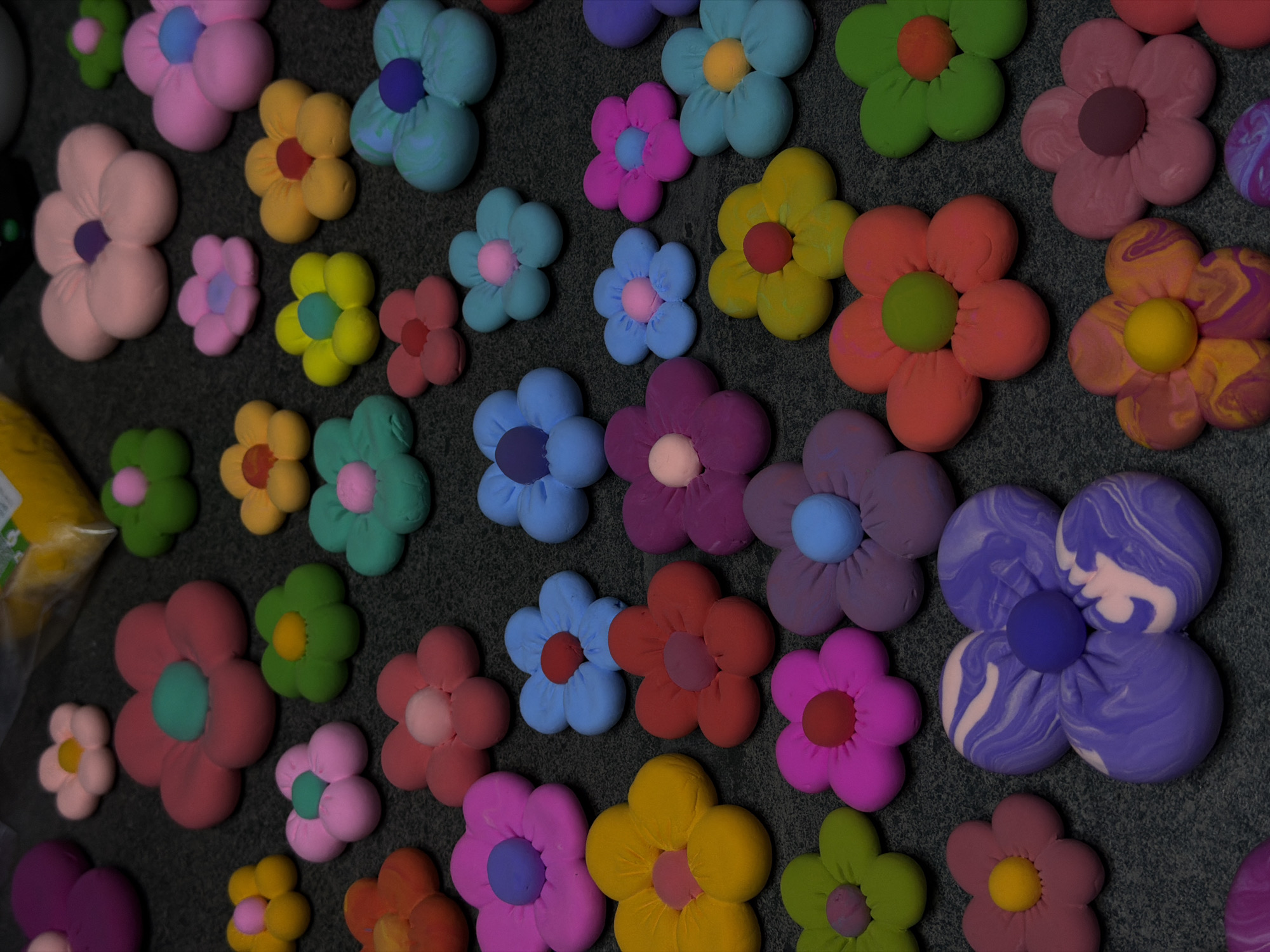 The image shows a collection of handmade clay flowers arranged on a dark gray surface. The flowers are of various colors including pink, yellow, green, blue, purple, and red, with contrasting colors used for the petals and the central part of the flower. Some flowers have marbled or swirled patterns. There are no people visible in the image. The location is not identifiable as Krasnoyarsk, Russia, based solely on the visual information provided.