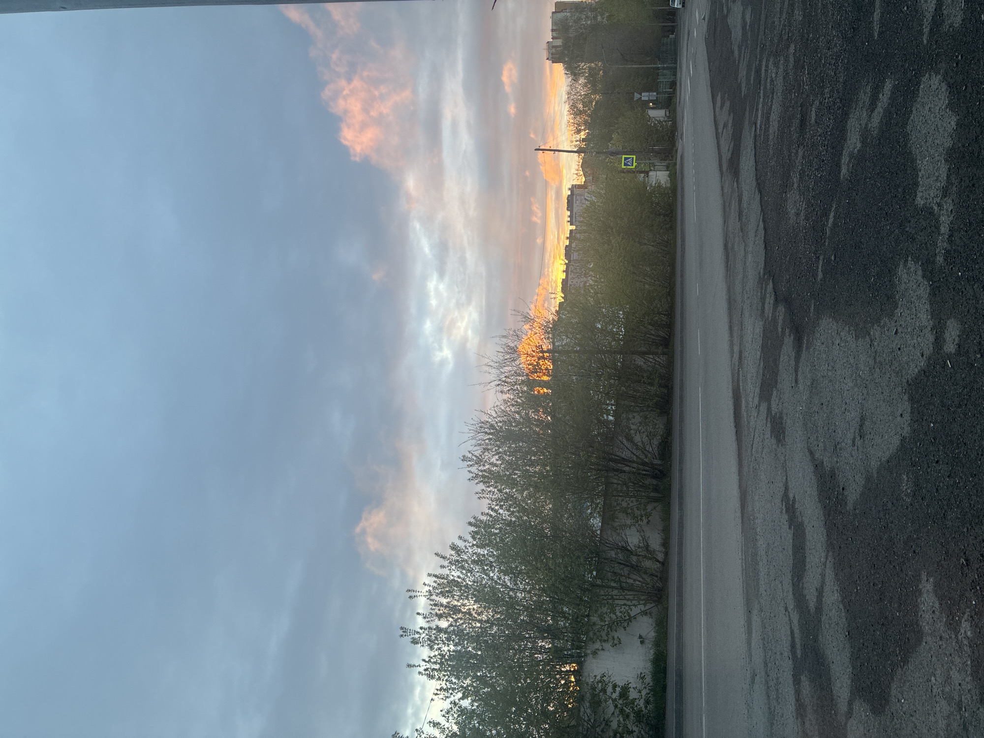 The image captures a view along a city street during a sunset. On the right side of the frame is the asphalt road, showing some signs of wetness. To the left of the road, a line of trees and bushes extends into the distance. In the background, buildings are visible, silhouetted against the bright sky. The sky dominates the upper portion of the image, featuring a mix of gray and orange-tinted clouds, indicating the setting sun. A pedestrian crossing sign is visible near the road. The location is Unknown, Unknown.