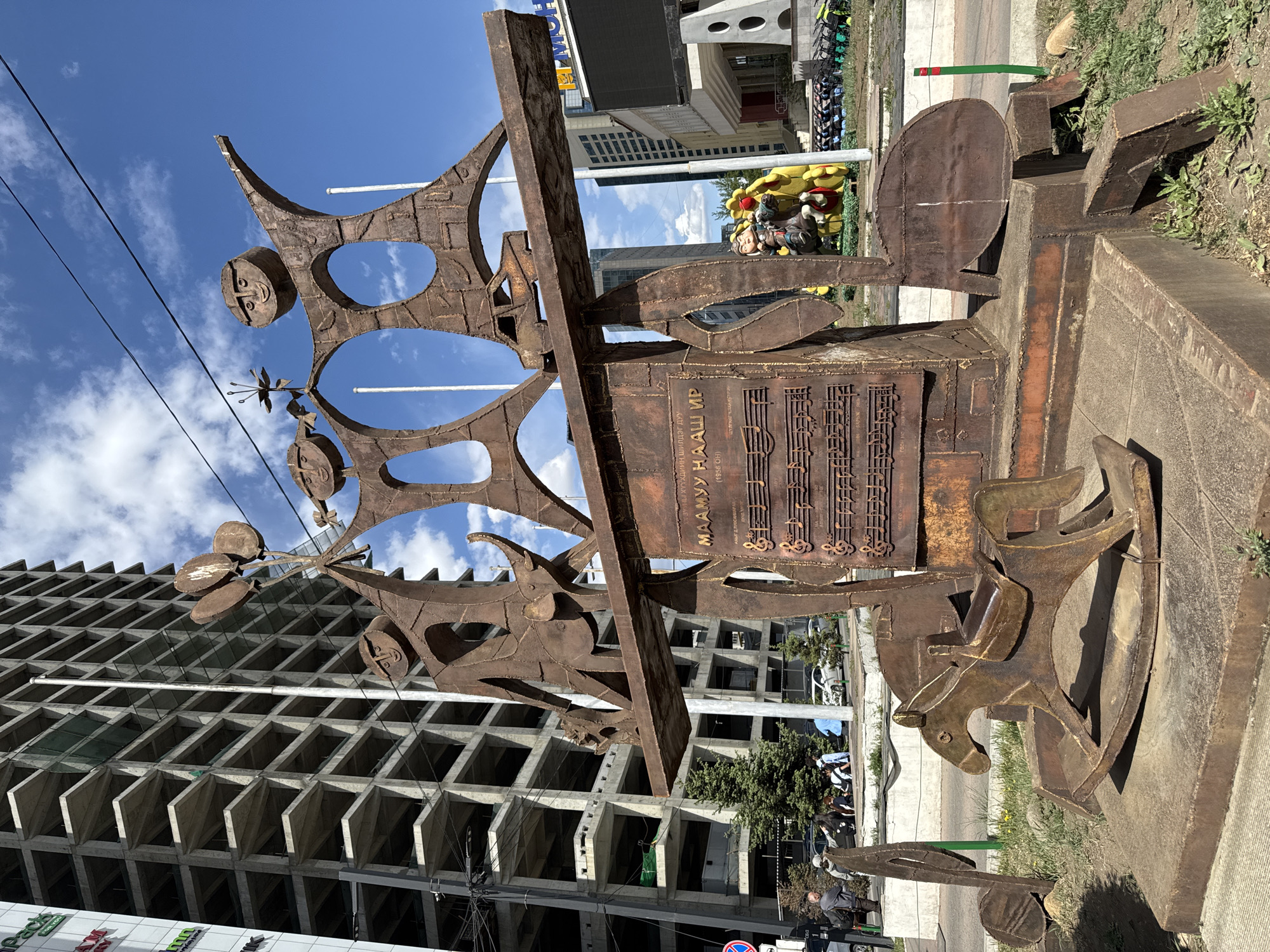 The image shows a corroded metal sculpture in Ulaanbaatar, Mongolia. The sculpture depicts stylized faces and musical notes, with Cyrillic script reading "МААМУУ НААШ ИР" (Maamuu Naash Ir) along with the text "(1956 OH)". In the background, a building with a geometric facade is visible, partially obscured by the sculpture. Additional city infrastructure elements like power lines and street signs are visible. Several passersby are walking on the sidewalk nearby.