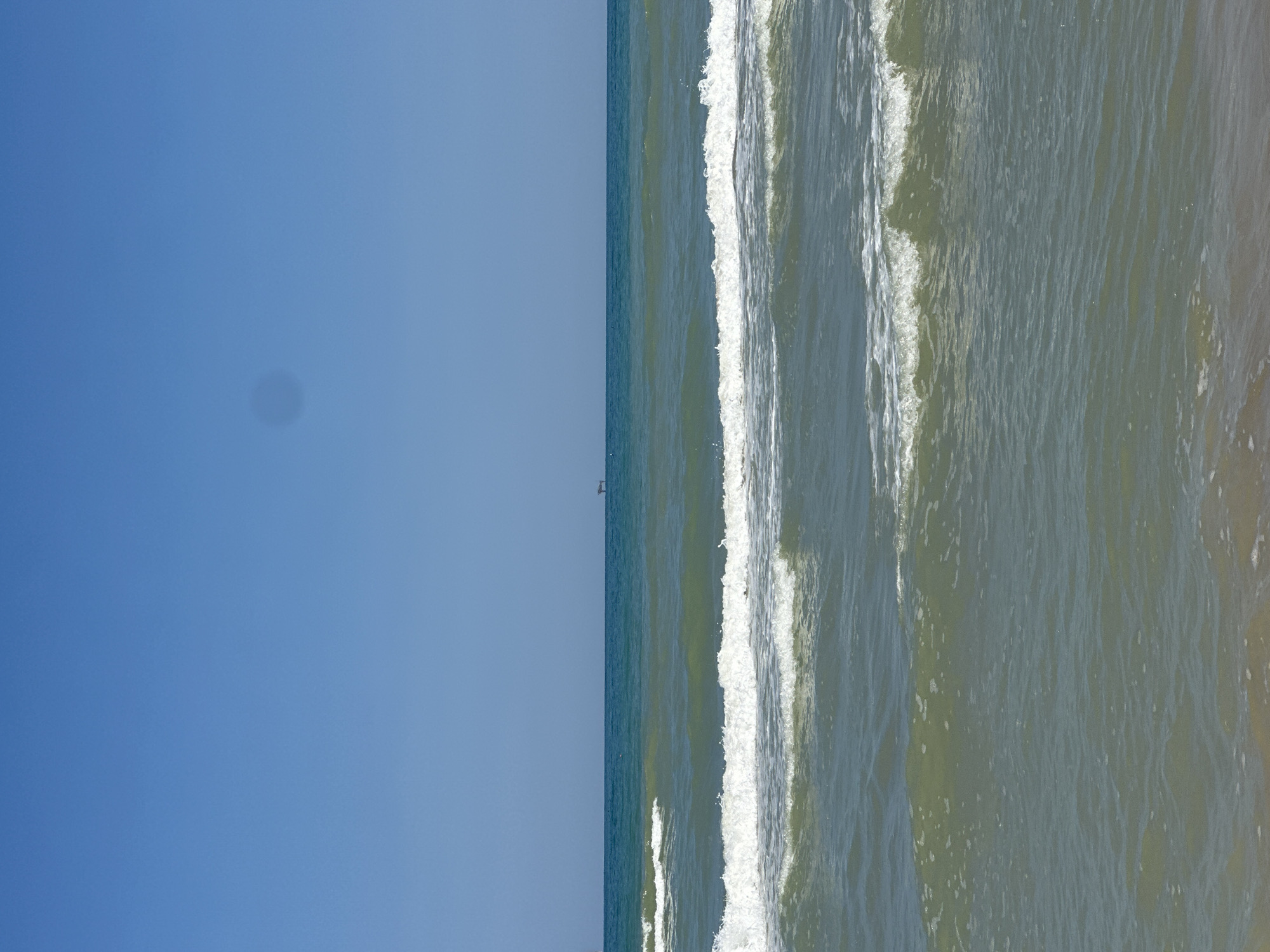The image shows a view of the Adriatic Sea from the shore in Pineto, Italy. The sky is a clear, vibrant blue, with a faint, circular smudge in the upper center of the image, possibly a lens flare or dust spot. The sea is a deep teal color, transitioning to a lighter, more greenish hue closer to the shore. Gentle waves with white foam crests are rolling in, with two prominent wave lines visible. In the distance, on the horizon, a small, dark, rectangular structure, possibly a buoy or a distant boat, is visible. The foreground shows a sandy beach with some wet sand and a few scattered white foam remnants from the receding waves. The overall scene depicts a calm, sunny day at the seaside.