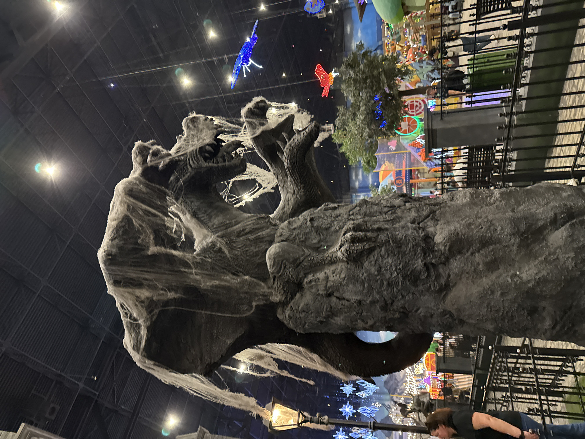 The image displays a large, dark, monster-like sculpture, possibly depicting a dragon or mythical beast, dominating the left and center foreground of an indoor scene. The sculpture is heavily draped in artificial white cobwebs, lending it a spooky, weathered appearance. Its mouth is open, revealing jagged forms, and its textured surface resembles rock or rough scales.
In the background, a brightly lit indoor entertainment complex unfolds. Suspended from the high, dark ceiling are several illuminated decorative figures, including a large blue butterfly-like light on the left and a red one to the right. Clusters of blue snowflake-shaped lights also hang in the mid-ground. A green tree, adorned with small blue lights, stands further back. Beyond this, colorful amusement park attractions, carousels, and other brightly lit structures create a festive atmosphere, separated by dark railings.
In the lower right corner, a person wearing a dark shirt and jeans is partially visible, appearing to observe the scene. A classic-style street lamp with an illuminated bulb is situated in the lower-middle portion of the image. The setting appears to be a large indoor space, such as an atrium of a shopping mall or an entertainment center in Moscow, characterized by high ceilings and varied decorative lighting, suggesting a themed event or permanent display. No visible text is present in the image.