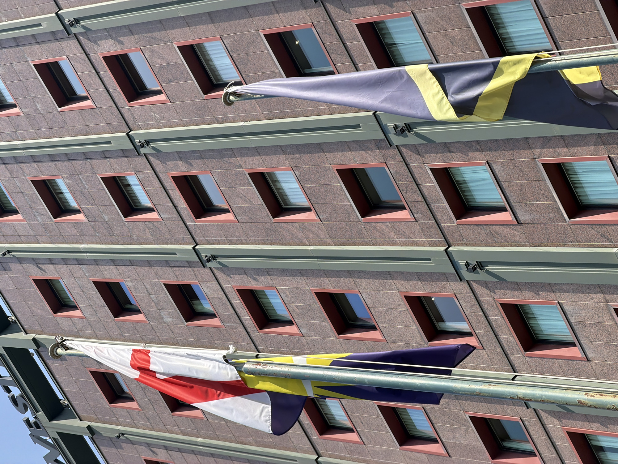 The image shows a close-up, low-angle view of the facade of a building, identified as the Westin Palace Hotel in Milan, Italy. The building's exterior is covered in dark red granite tiles with a slightly textured finish. The windows are recessed and have a red frame, and appear to be closed with sheer white curtains visible inside. Two flags are prominently displayed on flagpoles in front of the building. The flag on the left is predominantly white with a red cross, likely representing England. The flag on the right is a dark blue and yellow striped flag, which appears to be the flag of Ukraine. The sky is clear and blue, indicating a sunny day. The perspective is such that the building's facade stretches upwards, with several floors of windows visible.