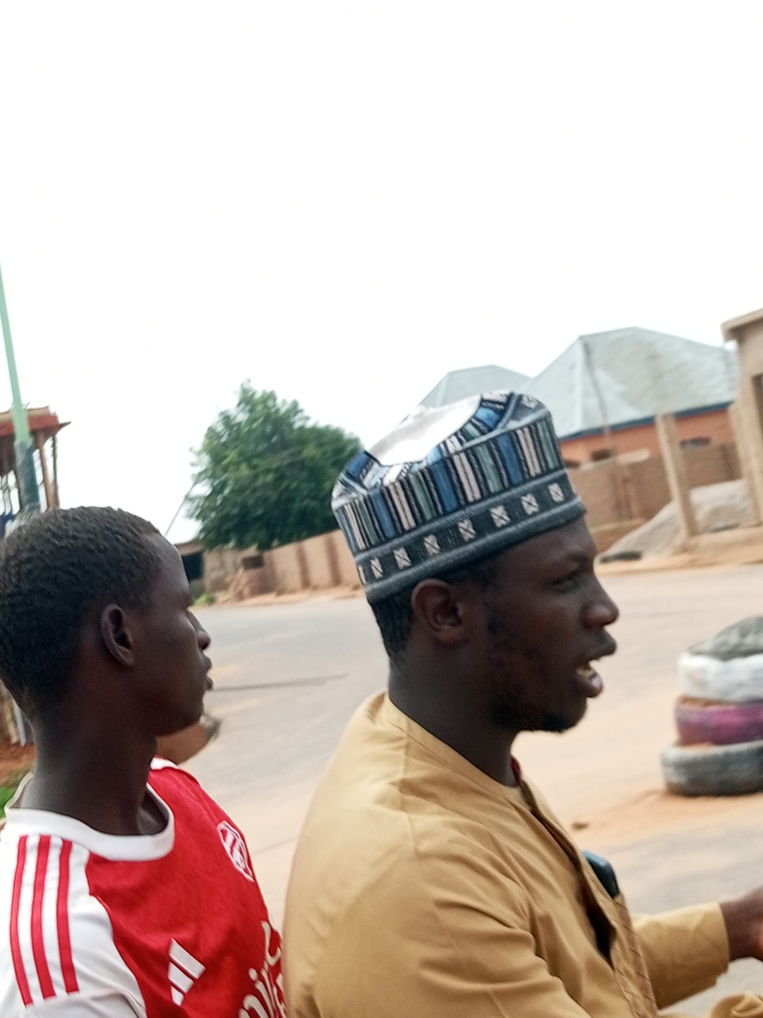 Two individuals, both male, are captured in profile against an overcast sky in what appears to be Tempure, Nigeria. The foreground is dominated by the upper bodies of the two men. On the left, a young man with dark skin and short, dark hair wears a red and white football jersey. His head is turned to the right, and his gaze is directed slightly downward. On the right, an adult male with dark skin wears a traditional cap with a patterned design of blue, white, and grey stripes and geometric shapes. He is also looking to the right, with his mouth slightly open as if speaking. He is dressed in a tan-colored garment. Behind them, the environment suggests an outdoor setting with a paved road and a dusty, sandy area. Several buildings with muted colors and angular roofs are visible in the middle ground, along with a lush green tree. In the far background, more structures are present. Stacked tires, with visible colored layers of purple and grey, are positioned in the right side of the frame. The overall atmosphere is casual and appears to capture a moment in everyday life in this Nigerian city.