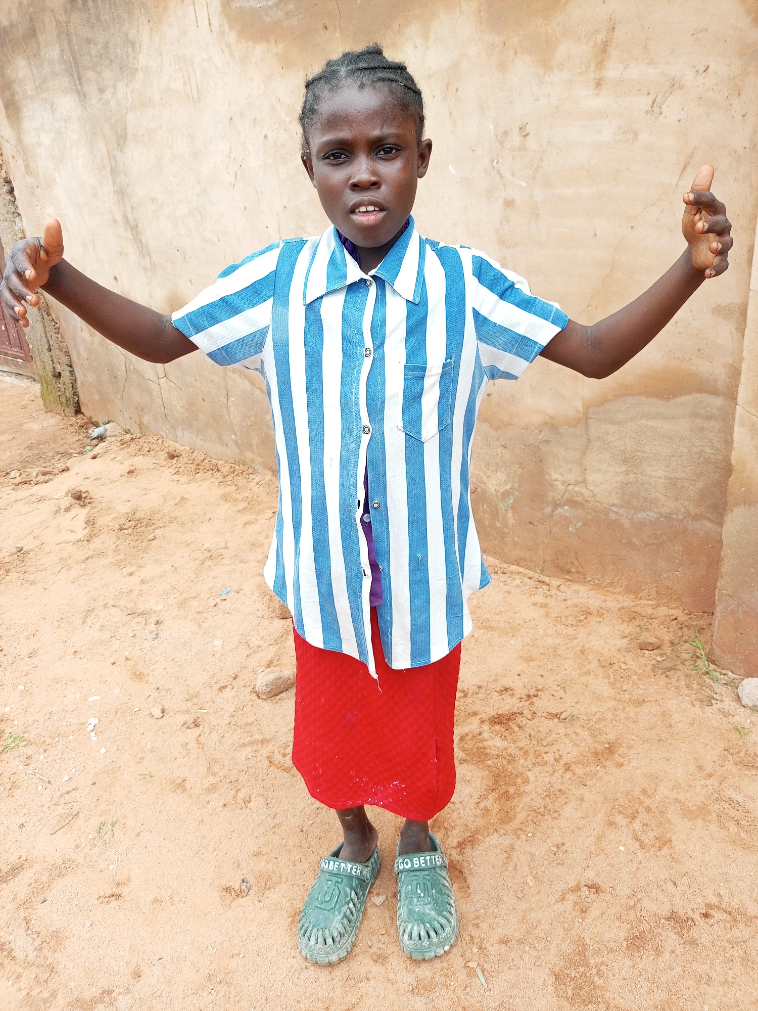 A young girl with dark skin and braided hair stands in an outdoor setting. She is wearing a blue and white striped short-sleeved collared shirt, a red skirt, and green clogs. The clogs have the word "GO BETTER" written on them. Her arms are outstretched to her sides, with her palms facing upwards. She is standing in what appears to be a dirt or sand-covered area with a rough, beige wall behind her. The wall has some texture and discoloration. The ground is sandy with some small rocks and debris. The overall scene suggests a simple, perhaps rural, environment. The location is identified as Tempure, Nigeria.