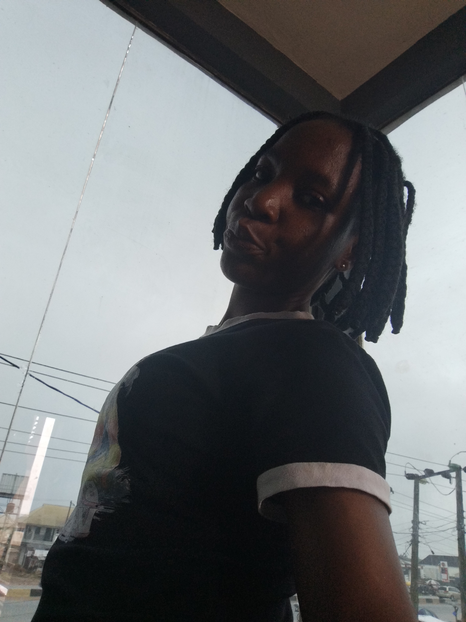 The image is a waist-up, slightly low-angle, indoor selfie taken by a person with dark skin and braided hair, looking upwards and to the left. They are wearing a black t-shirt with a light-colored trim around the collar and sleeve openings. The t-shirt has a graphic design on the left side, featuring faded colorful imagery that appears to be a depiction of a face or figure. The background shows a large window with a reflection of the sky, and beyond the window, a glimpse of a city scene with buildings, power lines, and utility poles, indicative of an urban environment. The general lighting suggests an overcast day. This scene is consistent with an interior location in Uselu, Nigeria, possibly a room with a large window overlooking a street.