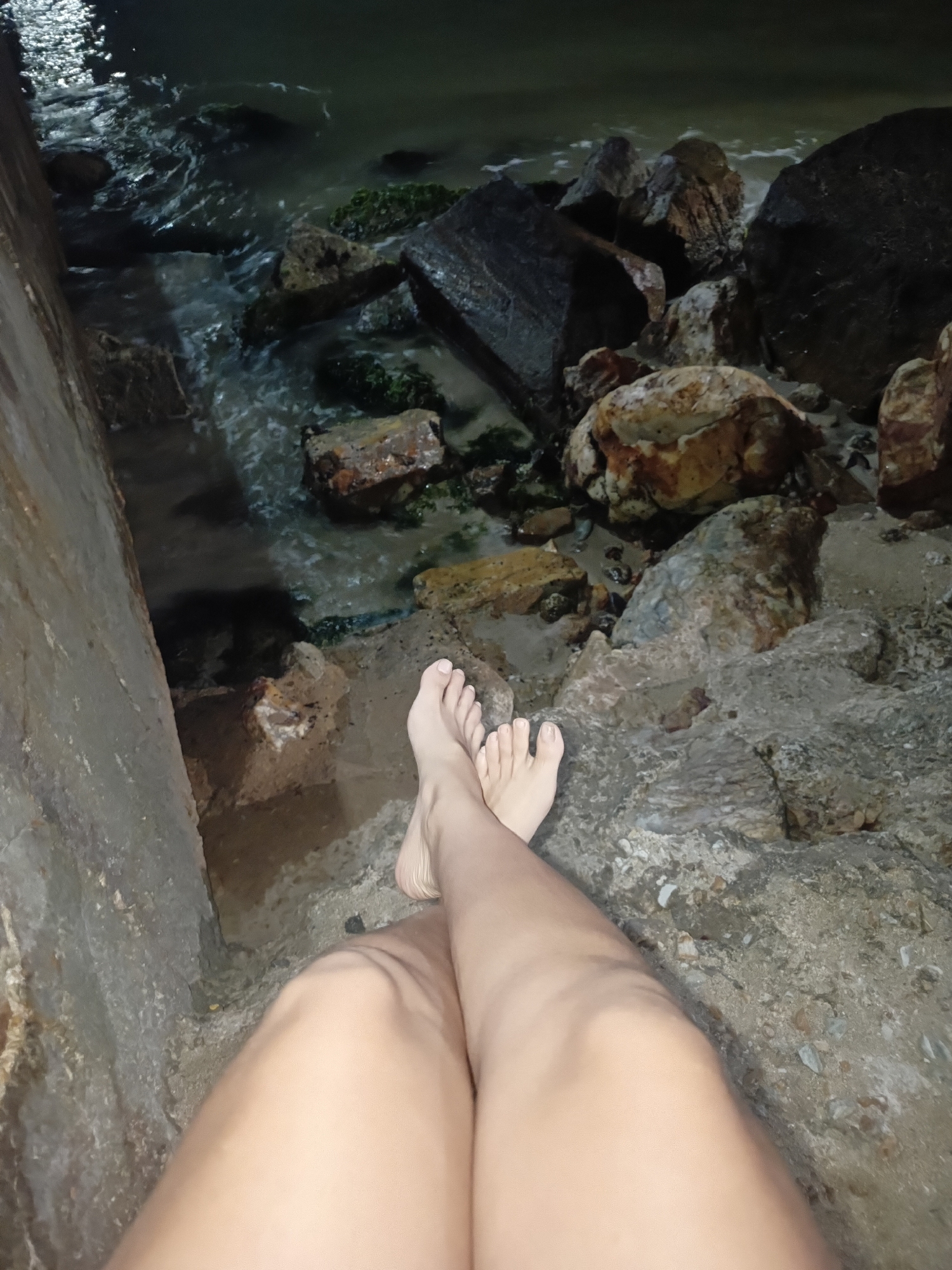 A low-light, first-person perspective photograph captures the bare lower legs and feet of an individual, crossed at the ankles, resting on a concrete surface. The scene is located at a rocky shoreline at night in Juan Griego, Venezuela. The concrete structure, possibly a seawall or embankment, occupies the left side of the frame and extends into the foreground. To the right and in the background, a cluster of various-sized rocks, some covered with dark green biological growth, borders dark water. The water exhibits subtle reflections of an unseen light source, indicating rippling movement. The ground adjacent to the concrete and beneath the rocks consists of light-colored sand and pebbles. The individual's skin tone is light to medium, and the toenails appear naturally colored or with a clear polish. No other persons, significant actions, or distinct objects are present within the frame.