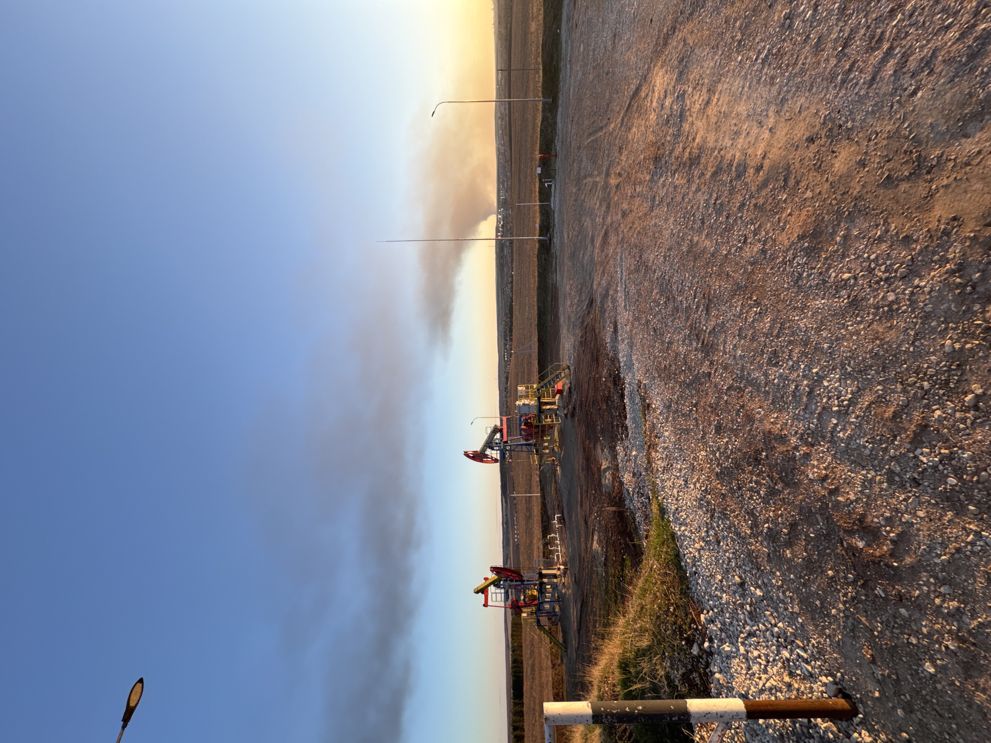 The image depicts an industrial scene near Kochkino, Russia. Several oil pump jacks are visible in the middle ground against a backdrop of open fields. Smoke or haze fills part of the sky. A gravel road is in the foreground with a barrier on its left. In the distance, some buildings and utility poles can be seen. The sky is blue with a gradient towards yellow near the horizon.