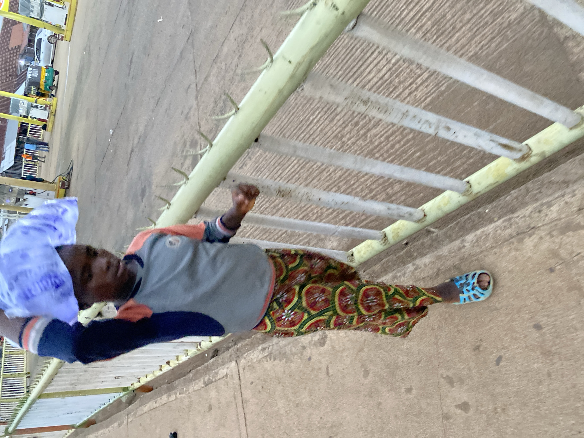 A man in Bukuru, Nigeria, is standing on a concrete surface next to a metal railing with spikes. He is wearing a gray and blue shirt, a patterned skirt, and blue sandals. A plastic bag is on his head. In the background, there is a building and a motorized tricycle.