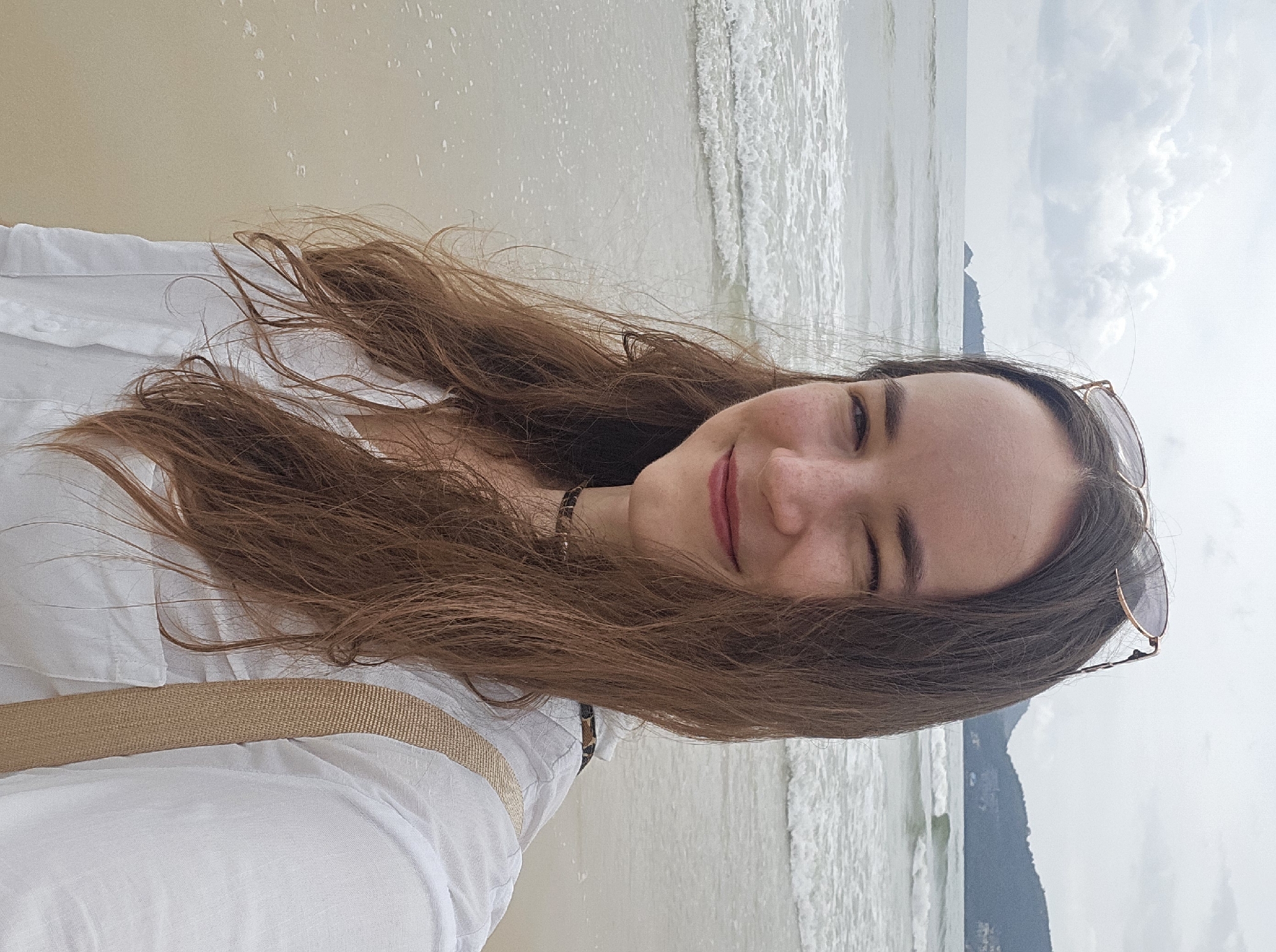 A young woman with long, brown, wavy hair winks at the camera while standing on a beach in Patong, Thailand. She is wearing a white linen shirt and sunglasses are perched on her head. A straw bag strap is visible on her shoulder. The ocean stretches out behind her, with gentle waves lapping at the shore. The sky is overcast with clouds.