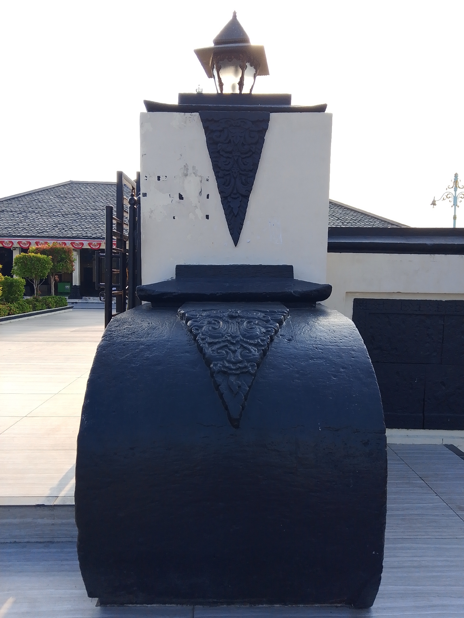 The image shows a close-up view of a large, dark, cylindrical structure with carved ornamentation at the front, positioned on a paved outdoor area. Above this structure, there is a white rectangular pillar with a V-shaped black design, topped by a traditional lamp. In the background, there are buildings with dark, textured roofs, and a glimpse of trees and a walkway. The overall scene suggests an outdoor public space, possibly a monument or part of a historical site. The location is identified as Demak, Indonesia, which is known for its historical Islamic architecture, particularly mosques and royal palaces. The style of the lamp and ornamentation hints at a cultural or historical context relevant to Indonesia.