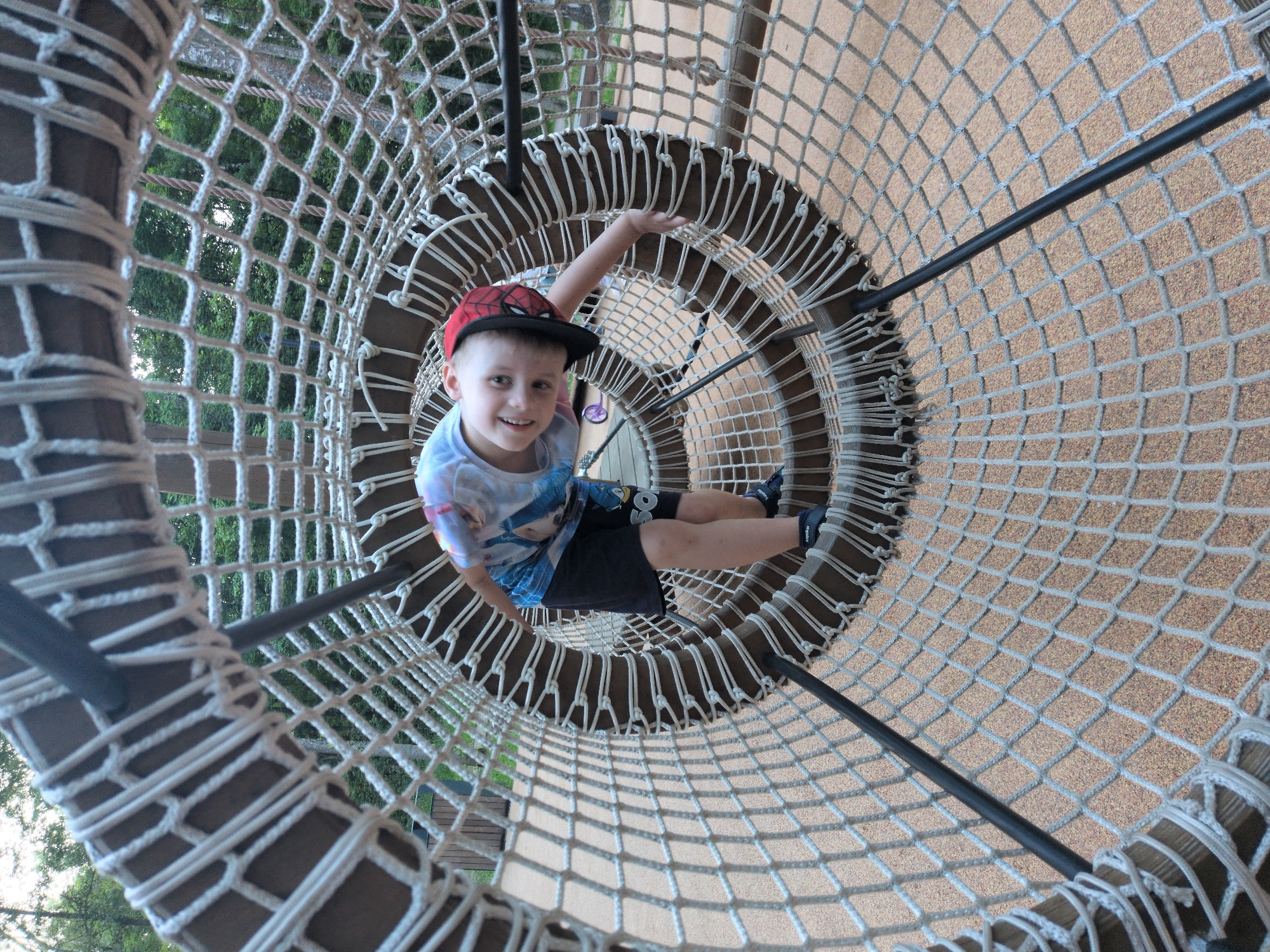 A young boy with blonde hair, wearing a red and black baseball cap with a Spider-Man emblem, a tie-dye t-shirt, and black shorts, is inside a cylindrical climbing net structure at an outdoor playground. He is smiling and looking towards the camera. His right arm is extended, and his left hand is gripping the inside of the net. The net is made of white rope and is supported by wooden beams and black poles. The ground beneath the net appears to be covered in a tan-colored safety surface. The background shows trees and foliage, suggesting a park or recreational area. The location is identified as the city of Pelikhovo, Russia.