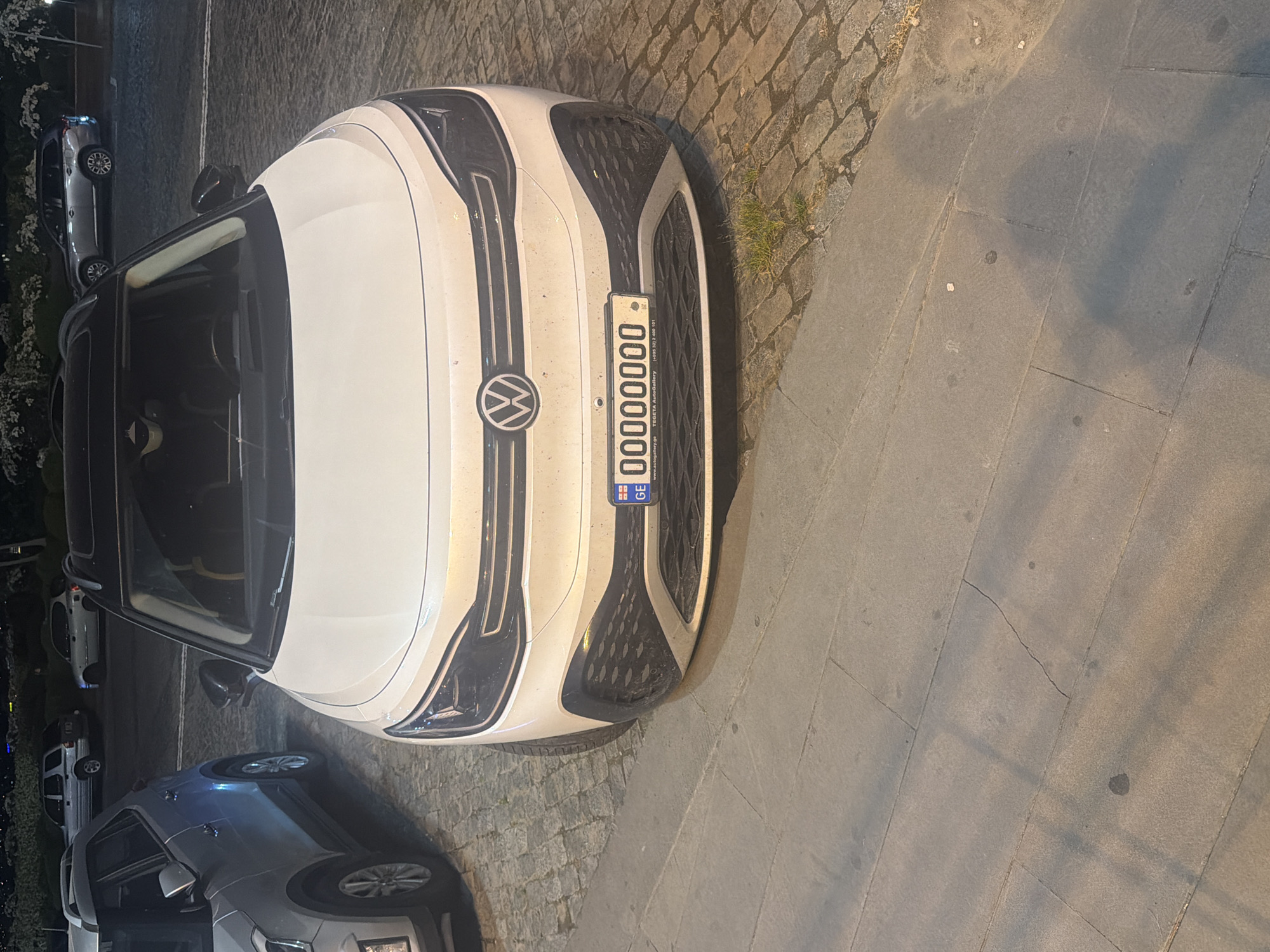 The image, captured at night or in the late evening, displays a white Volkswagen car, possibly a hatchback or small SUV, parked on a street. The car's front is prominently featured in the center, rotated 90 degrees clockwise within the frame, making it appear as if it's facing right. Its headlights are off, but the vehicle is illuminated by ambient light, highlighting its white body and black grille details. The circular Volkswagen logo is visible on the car's grille.
A Georgian license plate is clearly affixed to the front of the car. It features the country code "GE" in a small blue square on the left, followed by the numerical sequence "00000000". Below this number, smaller text provides contact information: "www.autobattery.ge", "TEGETA AutoGallery", and a phone number "+(995) 32 2 400 161".
The ground to what would be the car's left (appearing at the top of the rotated image) is a cobblestone surface, with small patches of grass growing between the stones. To the car's right (appearing at the bottom of the rotated image) is a flat, grey paved sidewalk. Other vehicles are partially visible, parked further down the street in both the background and foreground, suggesting a row of parked cars. Dark foliage or bushes are present in the far background. No people are visible, and no specific activity is taking place beyond the cars being parked. The scene depicts a quiet urban or suburban street environment.