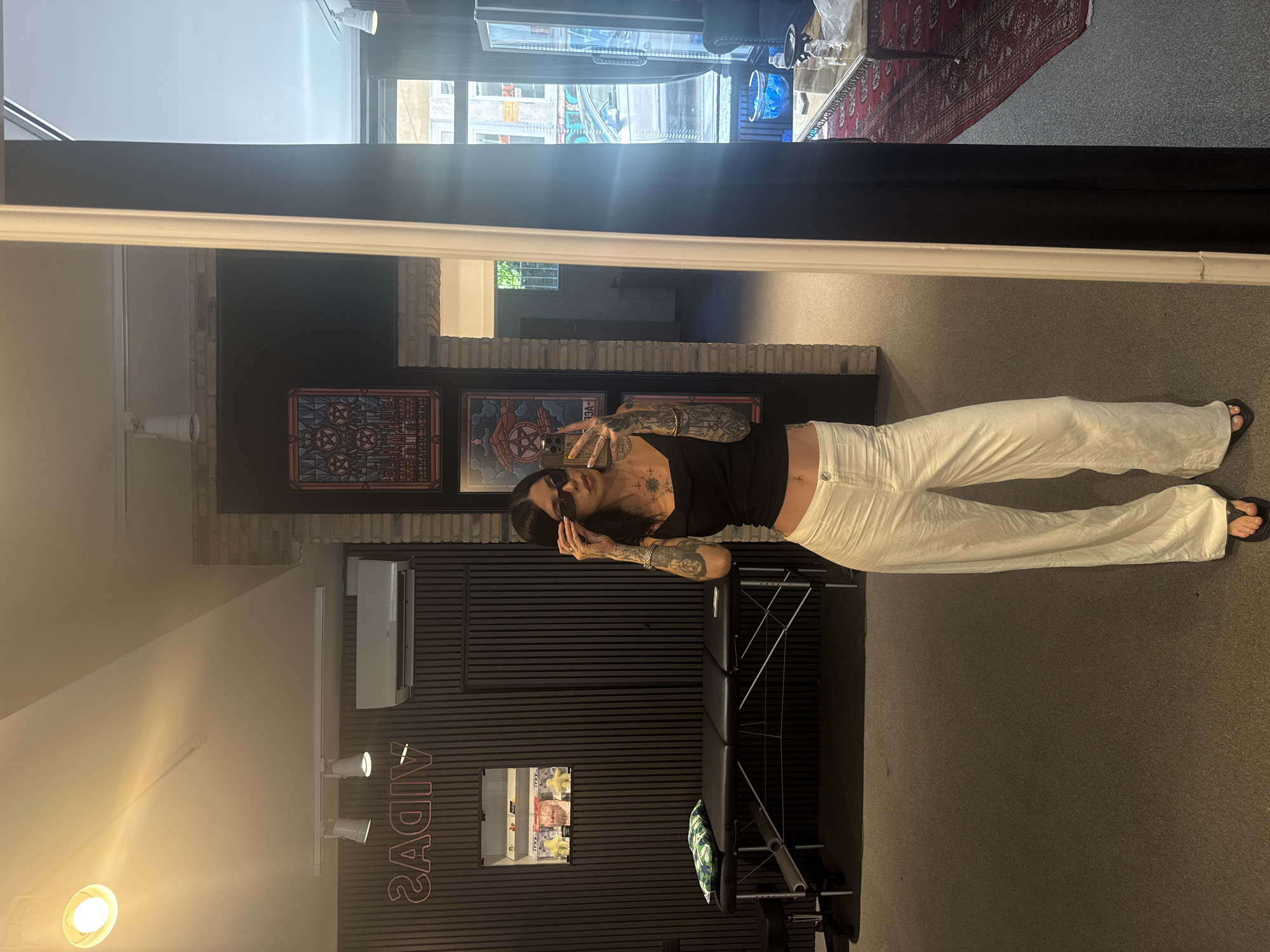 The image shows a woman in a tattoo studio, likely located in Sofia, Bulgaria based on the "Vidas" neon sign, a popular tattoo studio in the city. She is wearing sunglasses, a black top, and white pants, and is posing in front of a mirror, with a tattoo chair behind her. The studio has dark walls, artwork displayed, and an air conditioning unit. The reflection in the mirror shows the street outside, and a rug on the floor.