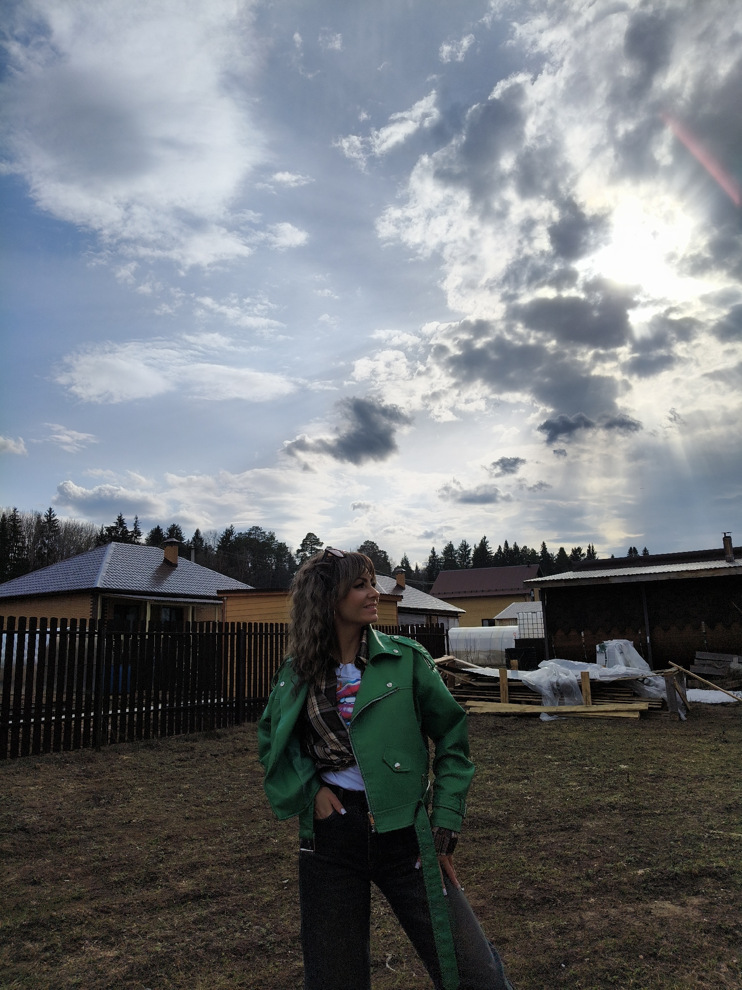 A person stands outdoors in a yard in what appears to be a rural area. The person is wearing a green jacket, jeans, and has curly brown hair. A plaid scarf can be seen around the neck. The yard is mostly brown grass, with a few scattered twigs and debris. In the background, there are several houses, some with wooden fences, and evergreen trees are visible in the distance. The sky is cloudy, with patches of blue and sunlight breaking through the clouds. The location could be Chaikovsky, Russia, given the user's prompt. Details visible in the background suggest a residential area on the outskirts of town, perhaps with seasonal homes or garden plots. There is construction material to the right of the frame with wood, plastic covering, and other assorted debris.