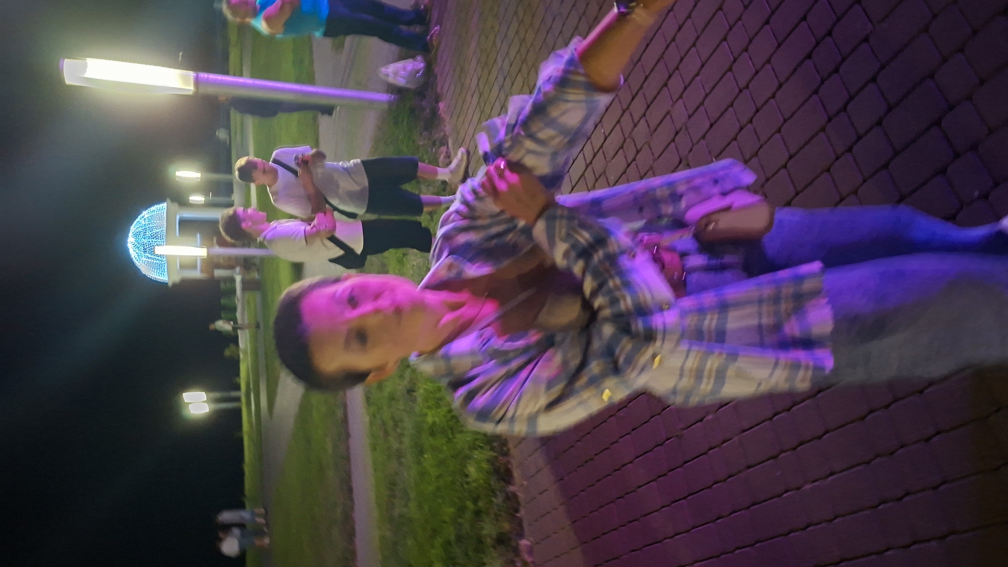 The image captures an outdoor scene at night, possibly in a city park or public space, with artificial lighting illuminating the area. In the foreground, a woman with short, dark hair and fair skin is the central subject. She is wearing a light-colored plaid shirt over a light-colored top and blue jeans. Her right arm is bent, and her hand is near her chest. A small, light brown purse is attached to her hip. The ground beneath her is paved with rectangular bricks, and the lighting casts purple and pink hues on the scene.
In the background, several other individuals are visible. Two young men are standing together, one in a white t-shirt and the other in a white polo shirt, both appearing to be engaged in conversation or looking at a phone. Further back, other figures are present, and a distinctive architectural structure with a domed roof adorned with blue lights is illuminated. Streetlights are also visible in the background, contributing to the nighttime illumination. The overall environment suggests a public gathering or event taking place after dark. The specific location in Myadzyel, Belarus, is not explicitly indicated by any visual cues within the image itself.