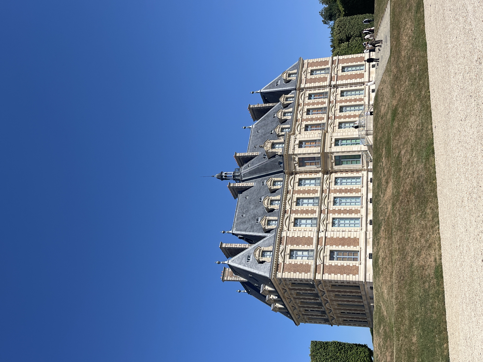 The image depicts the north facade of the Château de Sceaux, located in the Parc de Sceaux in Sceaux, France. The chateau is a large, ornate building with a mansard roof, intricate brickwork, and numerous windows. It stands on a gently sloping grassy hill.
In the foreground, a path of light-colored gravel leads towards the chateau. The middle ground is occupied by the grassy slope. The chateau itself is the primary subject, detailed with its architectural features.
On the steps leading up to the chateau, a group of approximately ten people is visible. They appear to be tourists, dressed in casual attire. Some are walking up the steps, while others are standing and observing the building. One individual in the foreground on the steps appears to be dressed in historical costume, possibly part of a reenactment or event.
To the left and right of the chateau, large, manicured hedges provide a geometric contrast to the building's architecture and the natural slope of the park. The sky is a clear, deep blue, indicating a sunny day. The overall scene conveys a sense of historical grandeur and public enjoyment of the park and its landmarks.