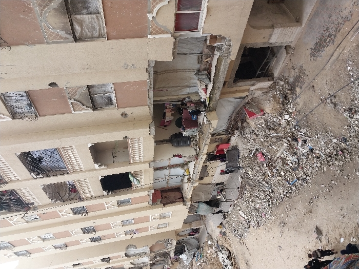 The image displays a street scene in the city of Khan Yunis, Palestinian Territory, characterized by severe structural damage to multi-story residential buildings. Multiple multi-story structures, primarily light-tan with sections of reddish-brown facade, line a dirt and rubble-filled thoroughfare. The lower two to three floors of these buildings exhibit significant damage, with large sections of exterior walls and interior partitions absent or collapsed, exposing interior rooms. Many windows are either broken, missing, or covered with plastic sheeting.
A wide expanse of concrete debris, rebar, and miscellaneous refuse covers the ground directly in front of the structures. A dirt path, marked by two parallel dark lines, traverses the debris field. Visible within the exposed interiors are items such as a large black water tank, red fabric hanging from a line, and a blue container. A wooden ladder is propped against a wall within a damaged section.
In the lower-left portion of the frame, approximately five to six individuals, dressed in dark attire, walk along the dirt path. One individual wears a dark head covering. Another lone figure is discernible near the exposed ground floor of a building further down the street.