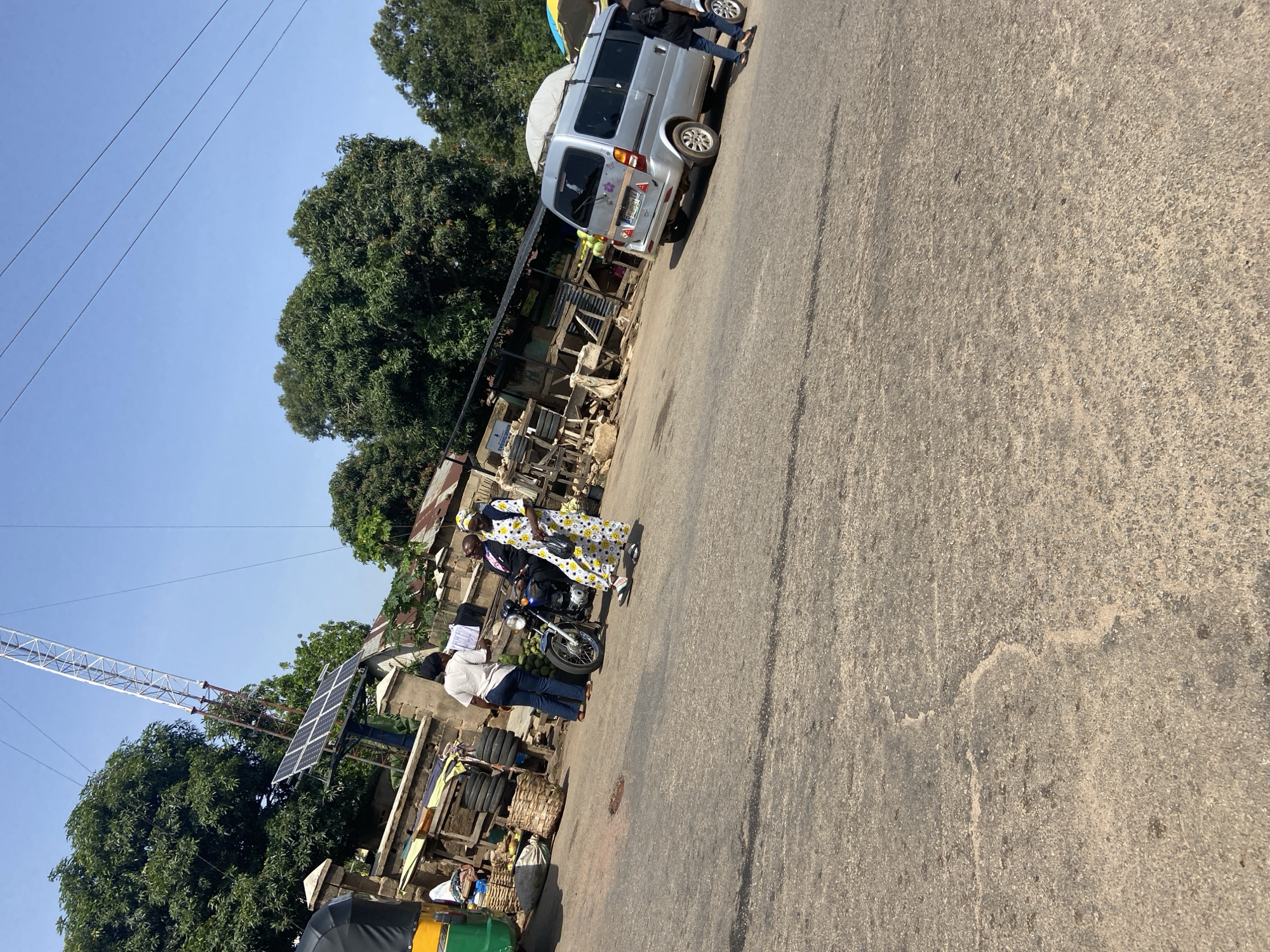 The image depicts a roadside scene in Jos, Nigeria, characterized by a paved road surface with visible cracks and patches, indicative of wear and tear. To the left, a green and yellow autorickshaw is parked, with a section of its black canopy visible. Adjacent to it, a roadside market stall displays an assortment of items, including stacked tires, woven baskets, and what appear to be produce, possibly fruits, arranged on the ground and within the baskets. A light-colored motorcycle is also present at the stall, with its kickstand down.
Several individuals are interacting around the stall and motorcycle. A person in a white t-shirt and blue jeans stands near the stall. Another individual, wearing a patterned dress with yellow flowers and a headscarf, is walking away from the motorcycle, carrying a dark bag. A person in a dark suit and tie, possibly talking to the person in the white t-shirt, is seated on the motorcycle. Another individual, partially obscured by the motorcycle, is also visible.
Further to the right, a silver minivan is parked on the road, facing away from the viewer. It has several passengers inside and a light-colored object resembling luggage or a tarp secured to its roof. Another person, wearing a dark t-shirt and jeans, walks towards the minivan, carrying a black backpack.
In the background, a large tower with what appears to be solar panels attached to its base stands prominently. Lush green trees with dense foliage surround the area, suggesting a locale with abundant vegetation. The sky is clear and blue, indicating fair weather conditions. The overall impression is a bustling, functional roadside market and transportation hub within the city.